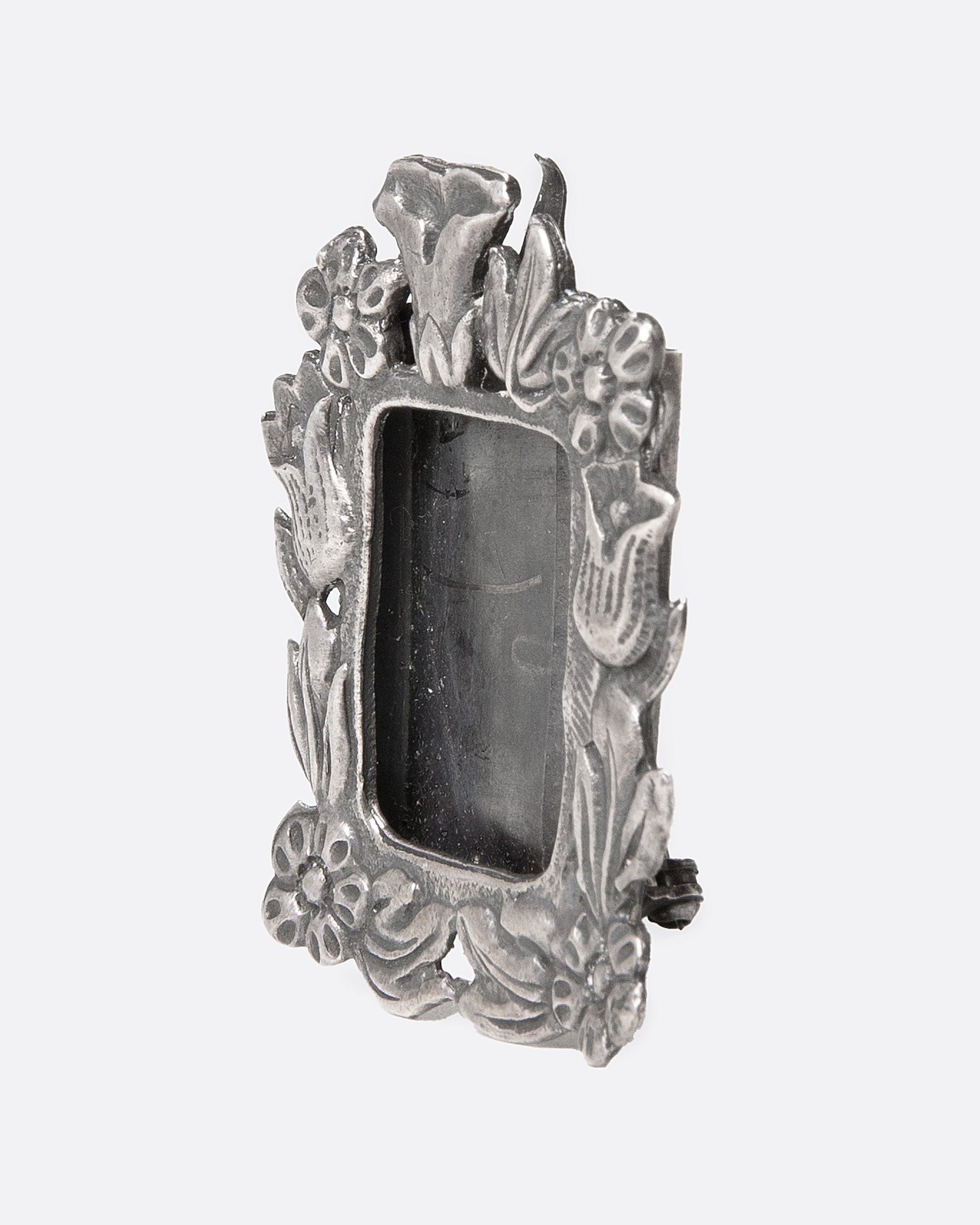 An ornate  silver picture frame brooch with a botanical motif around a rectangular opening, shown from the side.