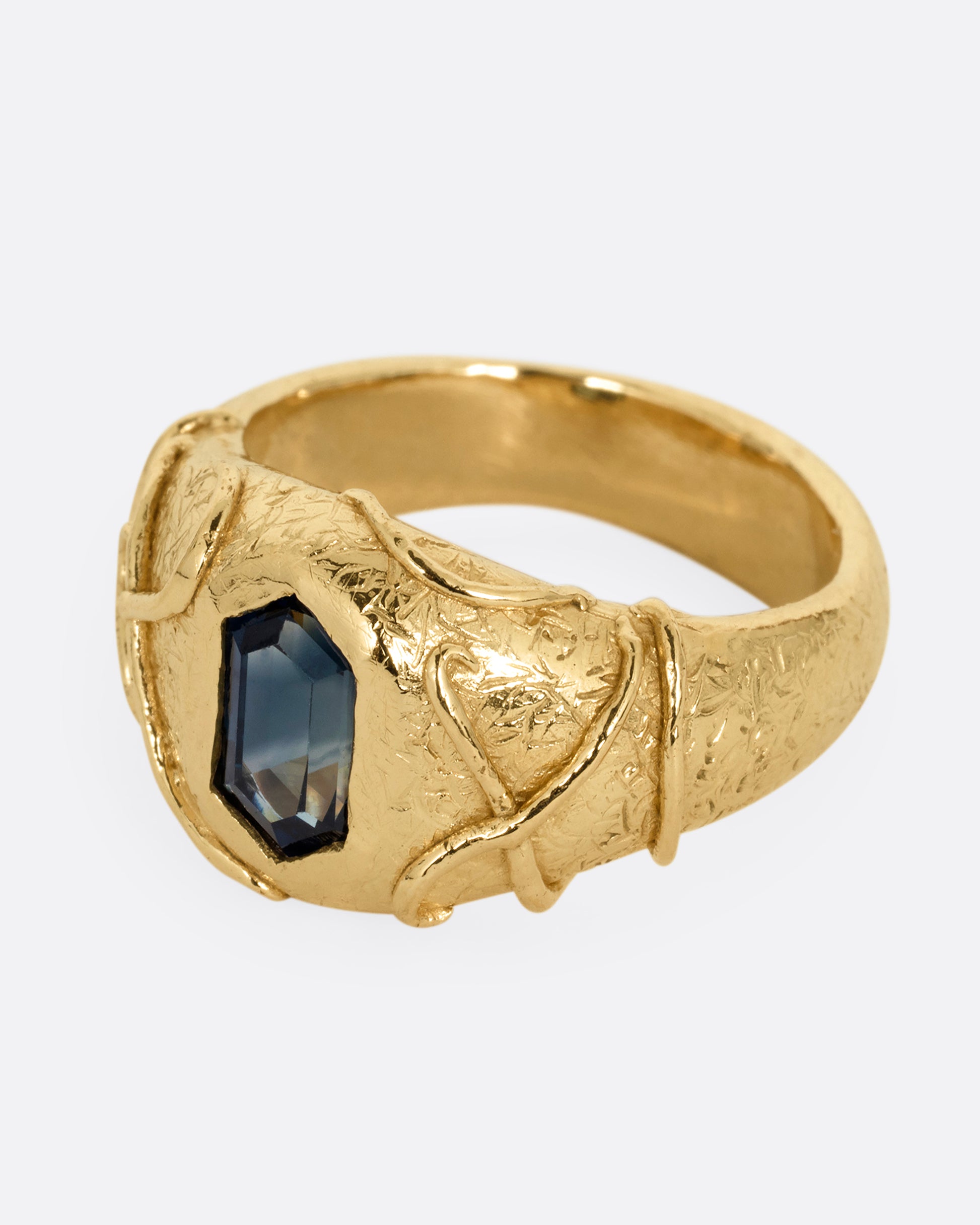 A yellow gold signet ring with a vine motif and a  hexagonal blue sapphire. Shown from the side.
