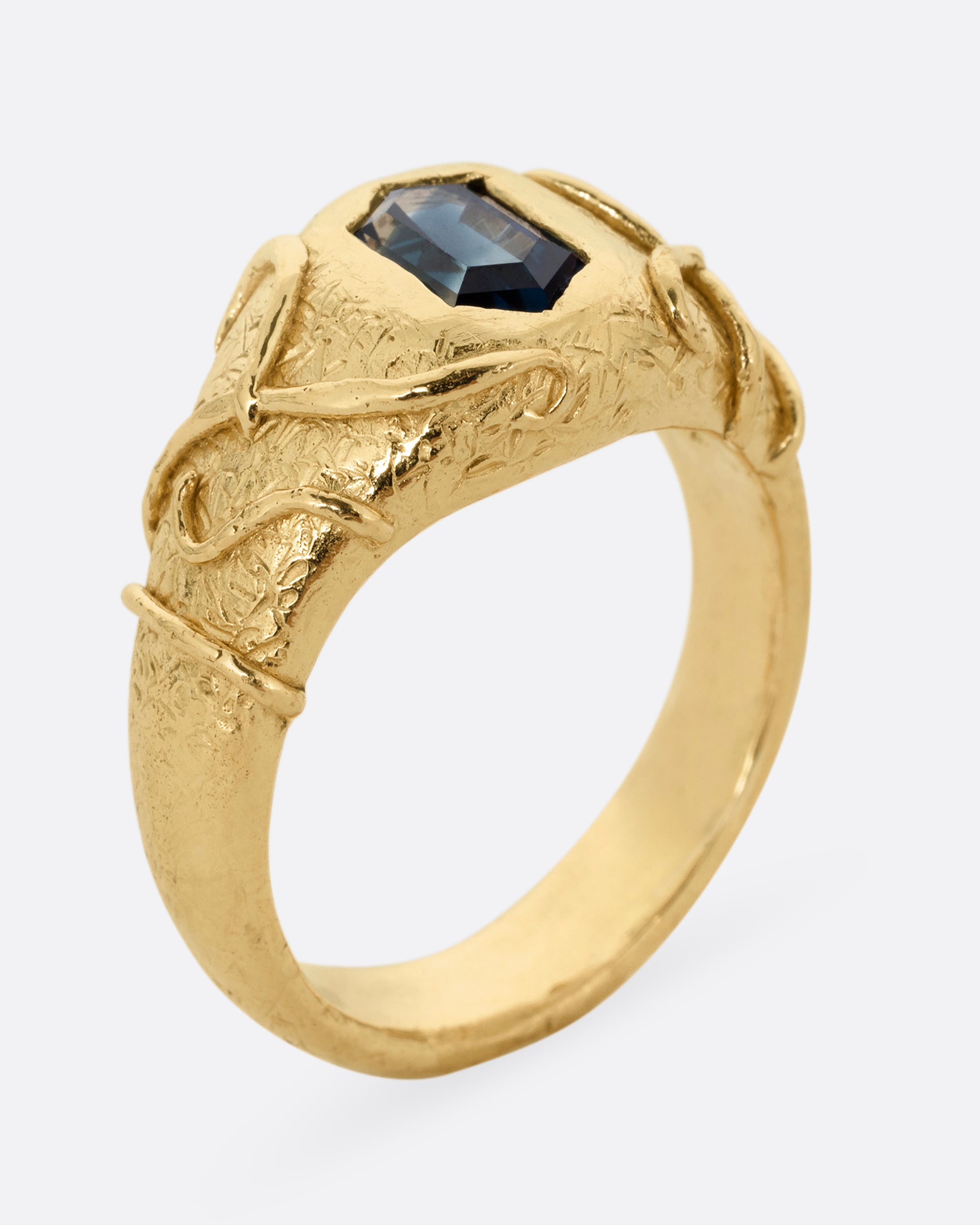 A yellow gold signet ring with a vine motif and a  hexagonal blue sapphire. Shown standing up from the side.