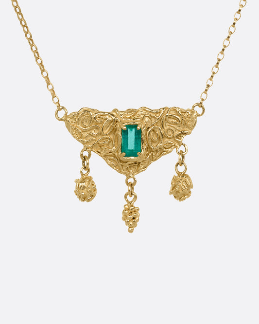 A yellow gold necklace with an emerald at the center of a sculptural pendant and three dangles- two blooming roses and a cluster of grapes. Shown from the front.
