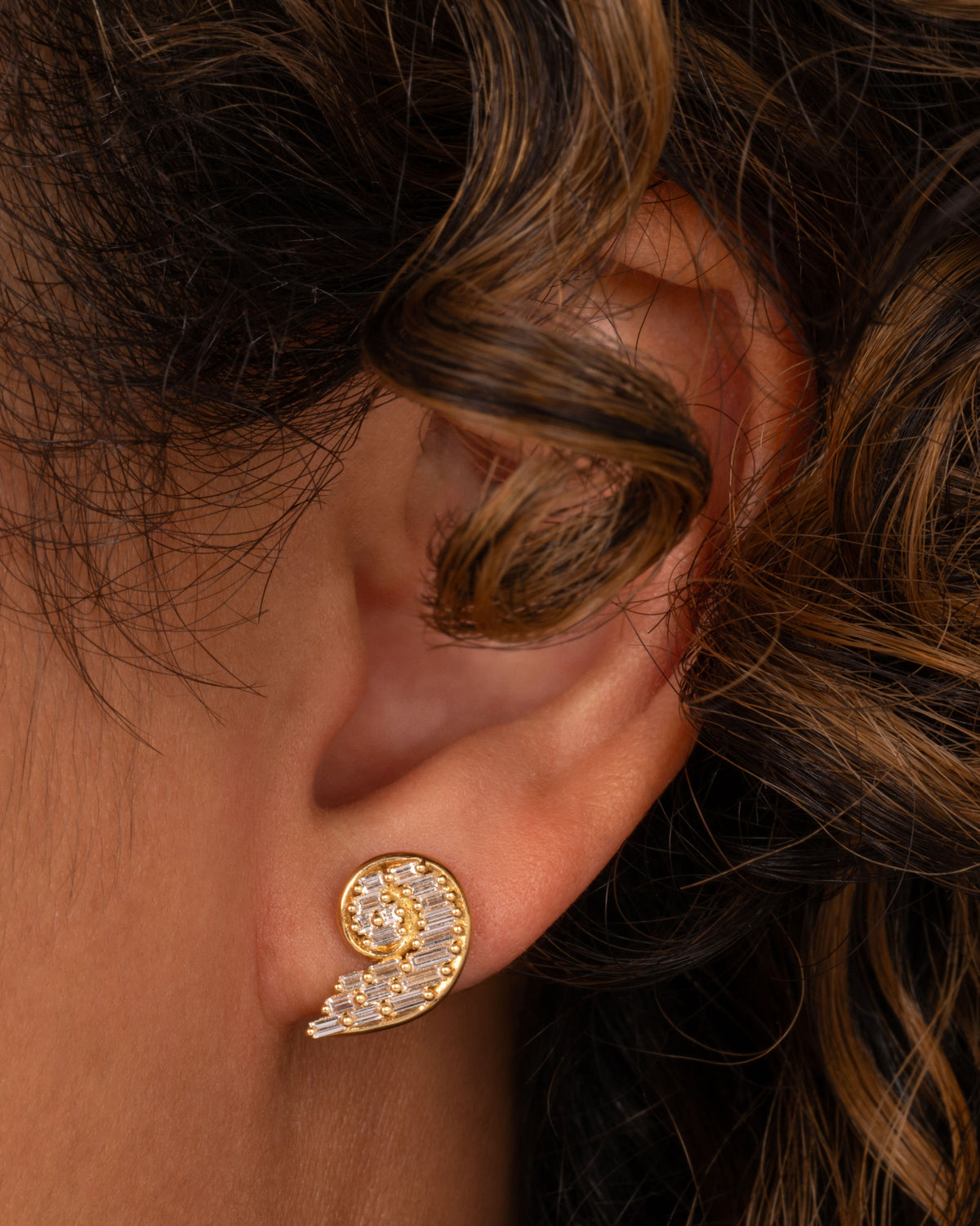 Artemer shell stud earring in yellow gold worn on an ear, a curved spiral shape set with baguette diamonds
