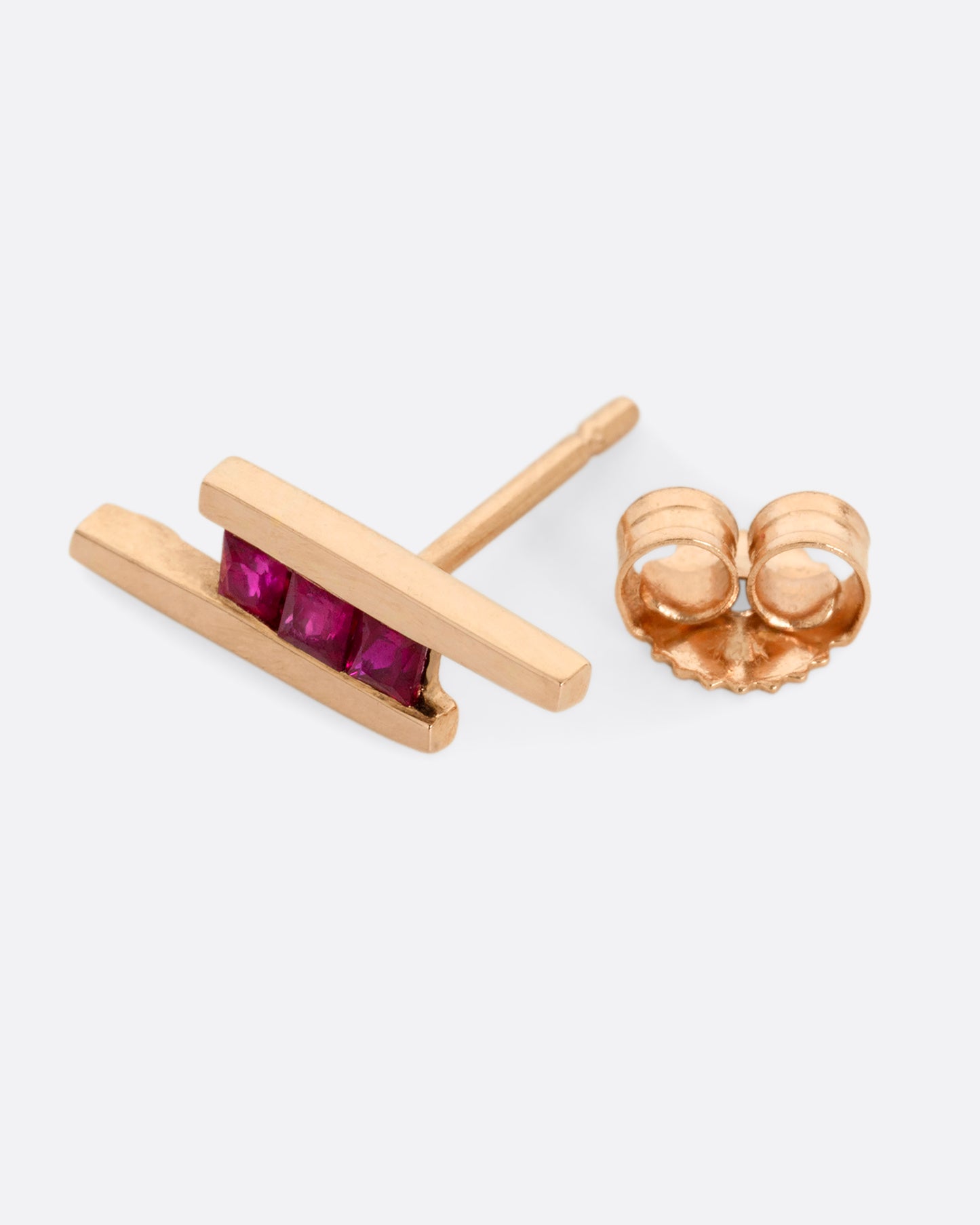 A rose gold stud earring with three rubies between two bars. Shown lying flat open.