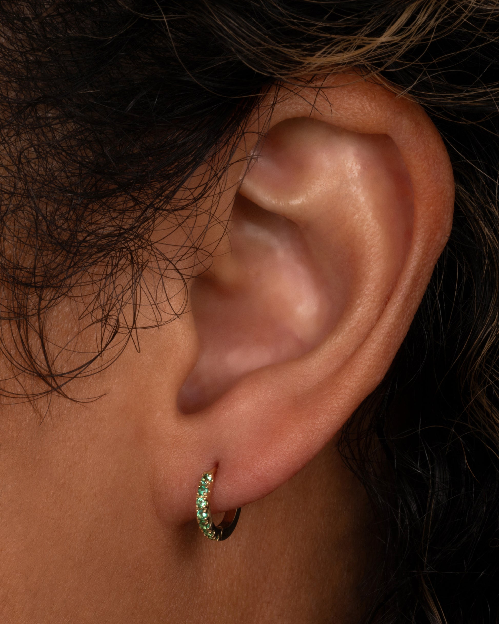 A yellow gold hinged hoop earring with pavé emeralds, shown on a person.