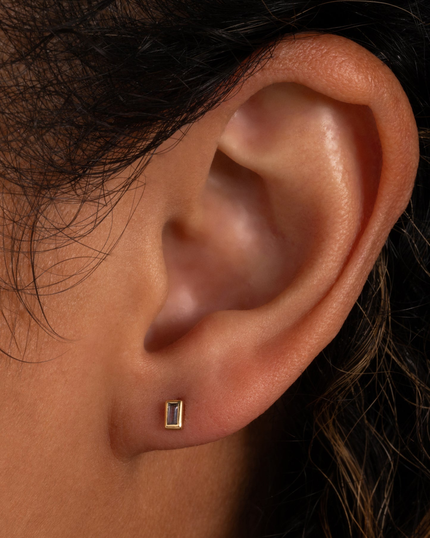 A yellow gold stud earring with a baguette cut lavender stone. Shown in an ear.