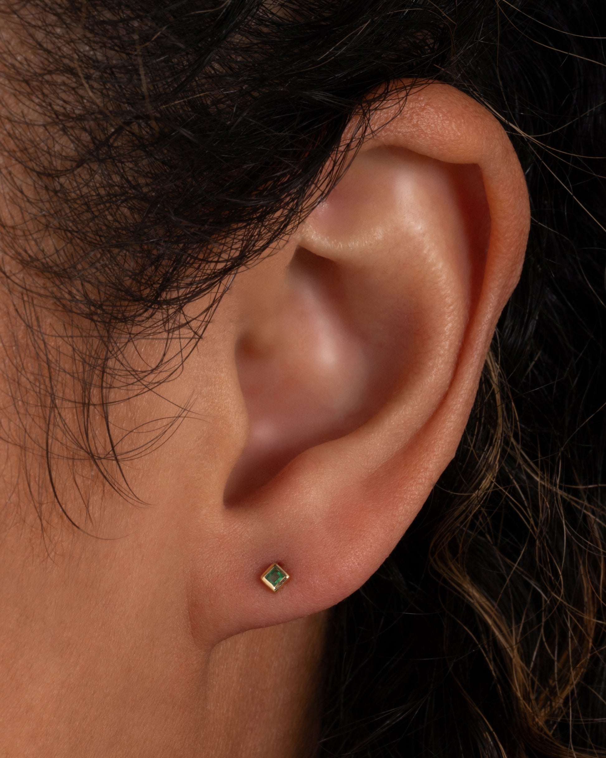 A yellow gold stud earring with a princess cut emerald. Shown in an ear.