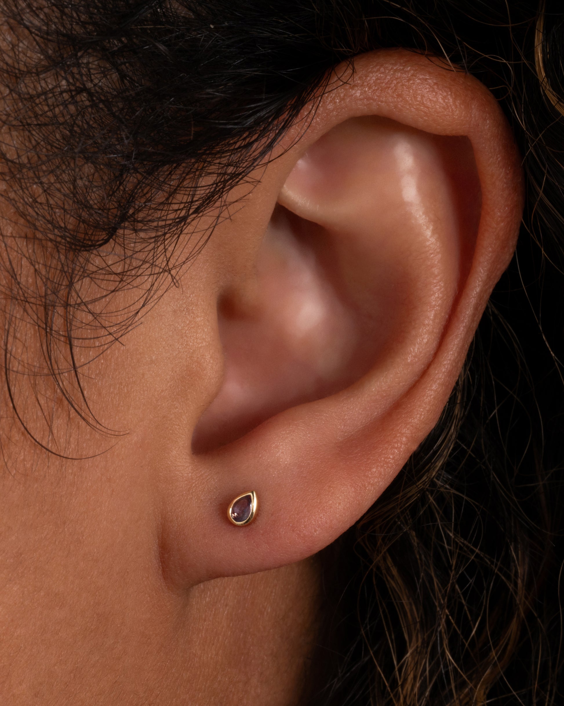 A yellow gold teardrop shaped bezel set tanzanite stud earring. Shown on a person.