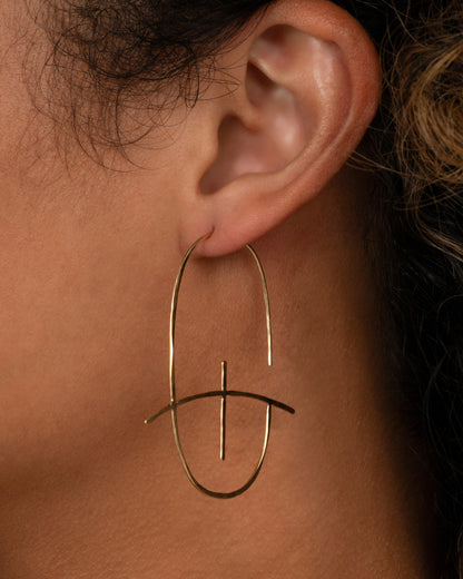 Christine Fail large Altar hoop earring in Fairmined 14k gold worn on an ear, a long hammered oval wire crossed by a curved bar
