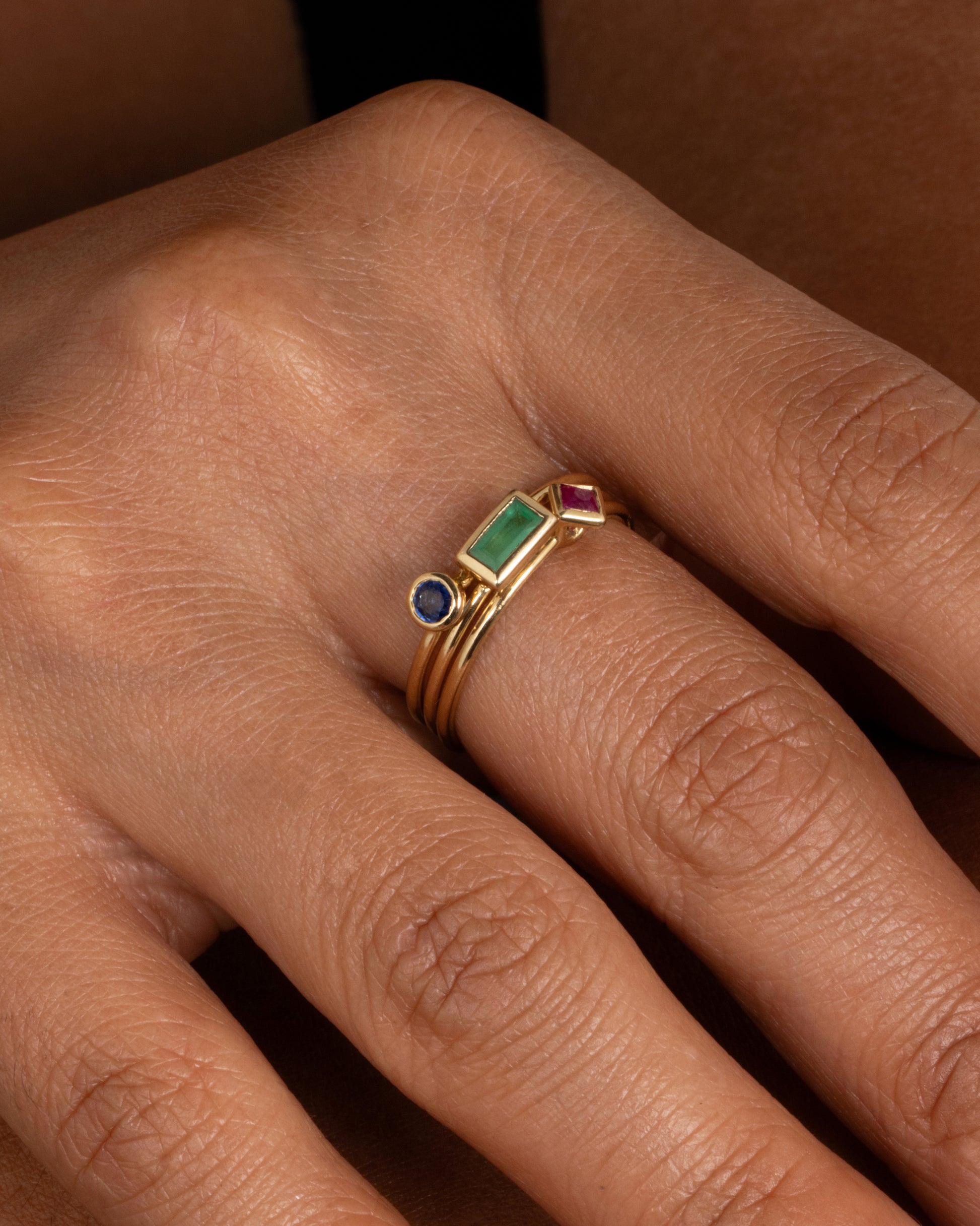 A stack of yellow gold rings- one with a round sapphire, one a rectangular emerald, and one a princess cut ruby. Shown on a hand.