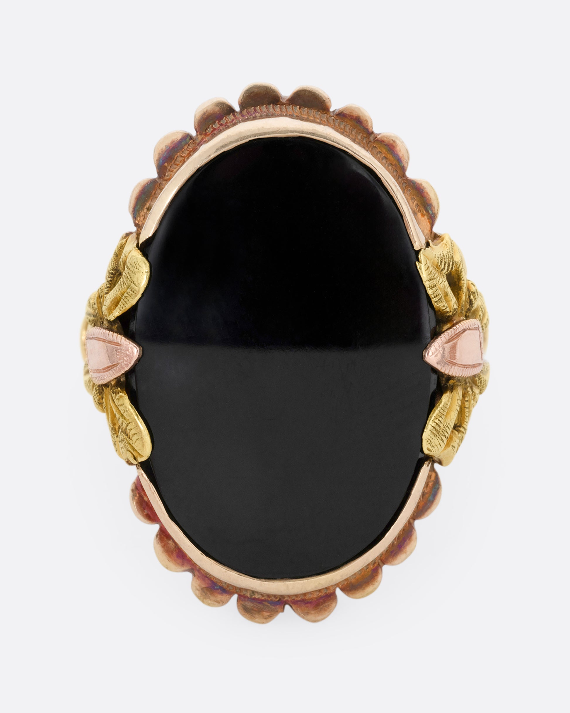 A yellow, rose, and green gold ring with a scalloped bezel, bow- topped shoulders, and an oval onyx. Shown from the front.
