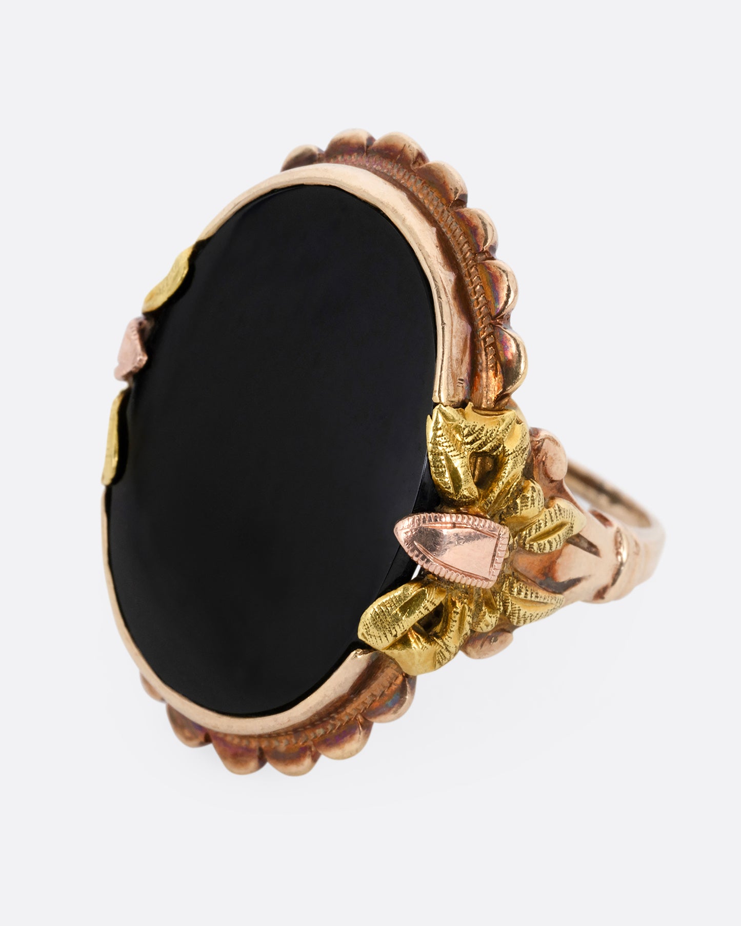 A yellow, rose, and green gold ring with a scalloped bezel, bow- topped shoulders, and an oval onyx. Shown from the side.