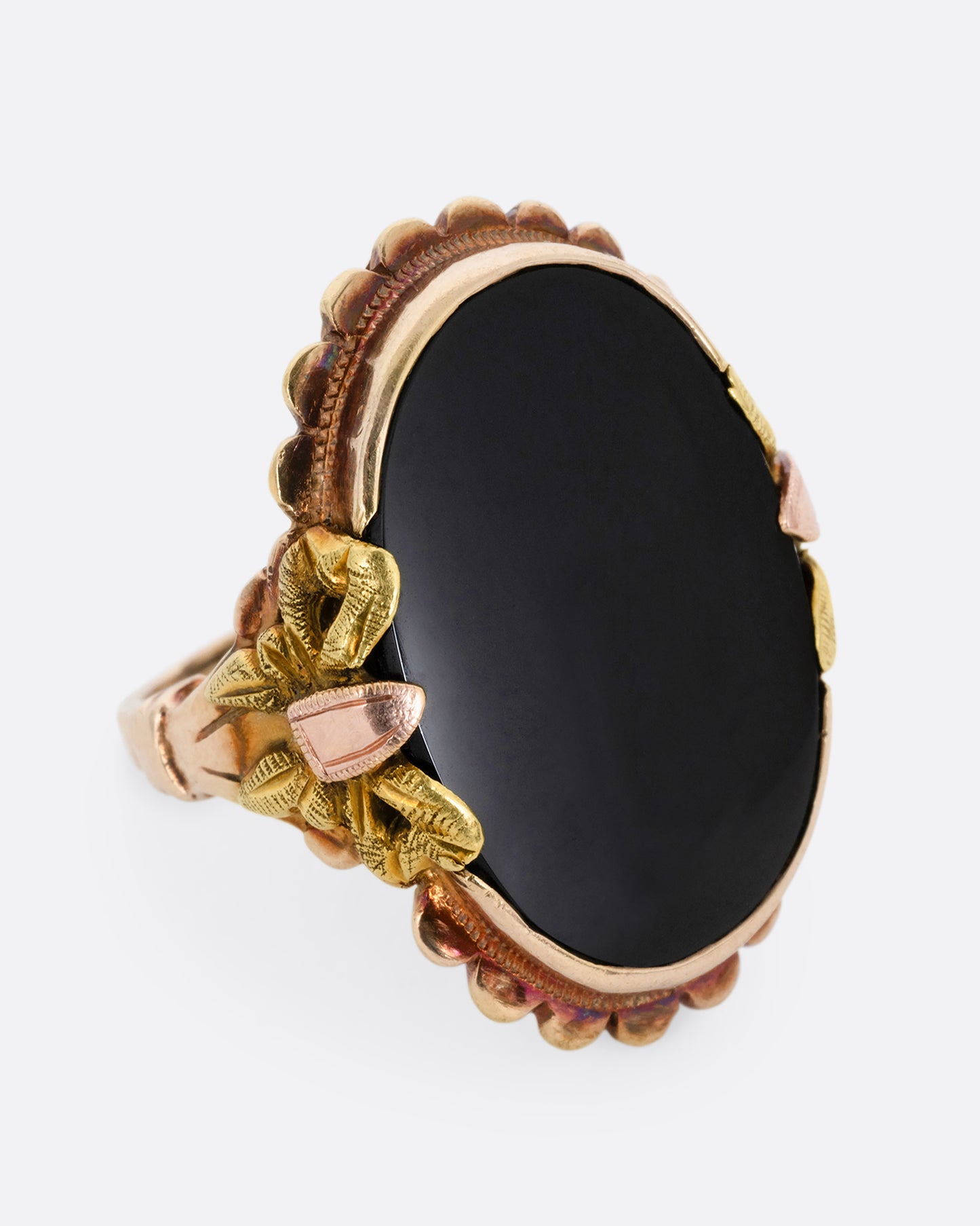 A yellow, rose, and green gold ring with a scalloped bezel, bow- topped shoulders, and an oval onyx. Shown from the side.