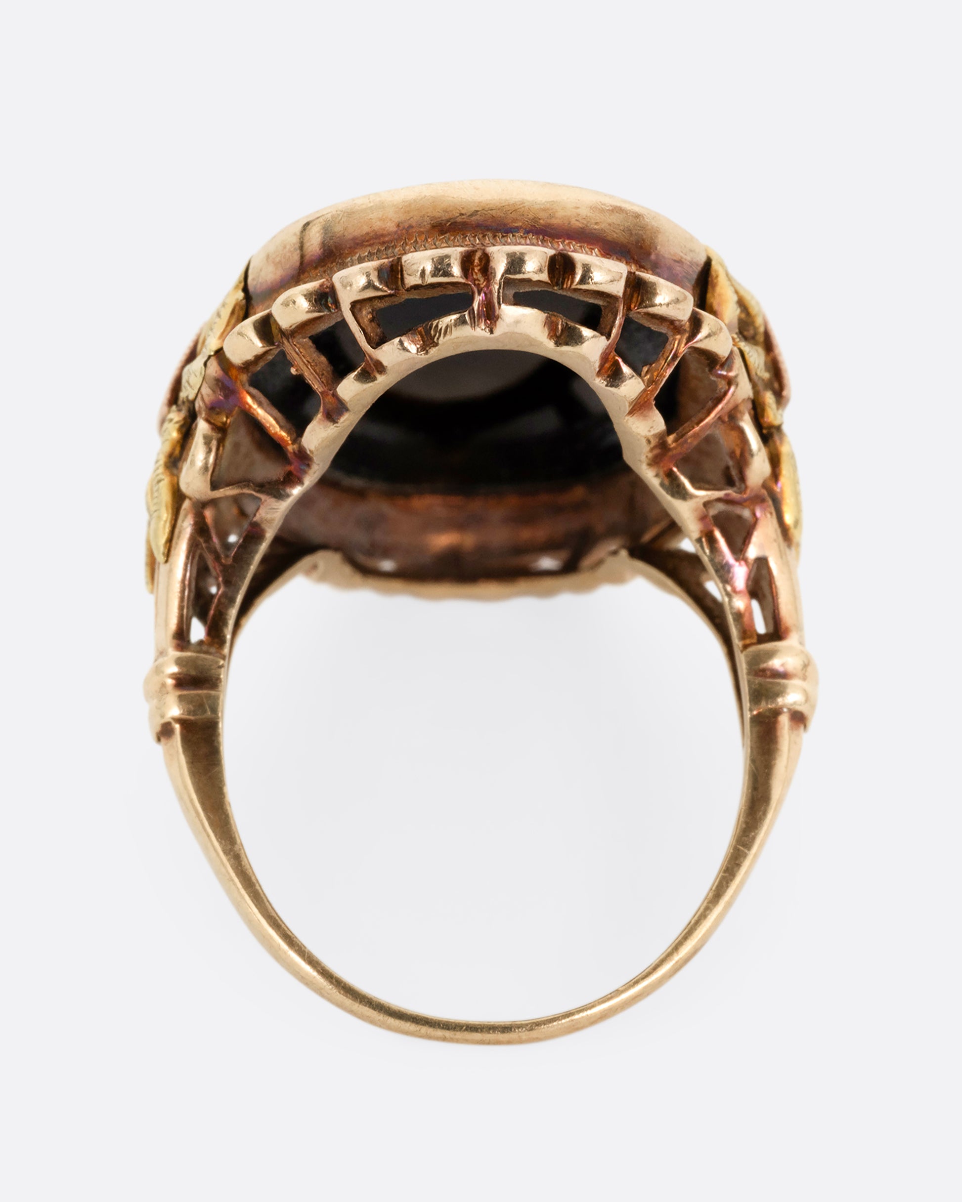 A yellow, rose, and green gold ring with a scalloped bezel, bow- topped shoulders, and an oval onyx. Shown from the back.