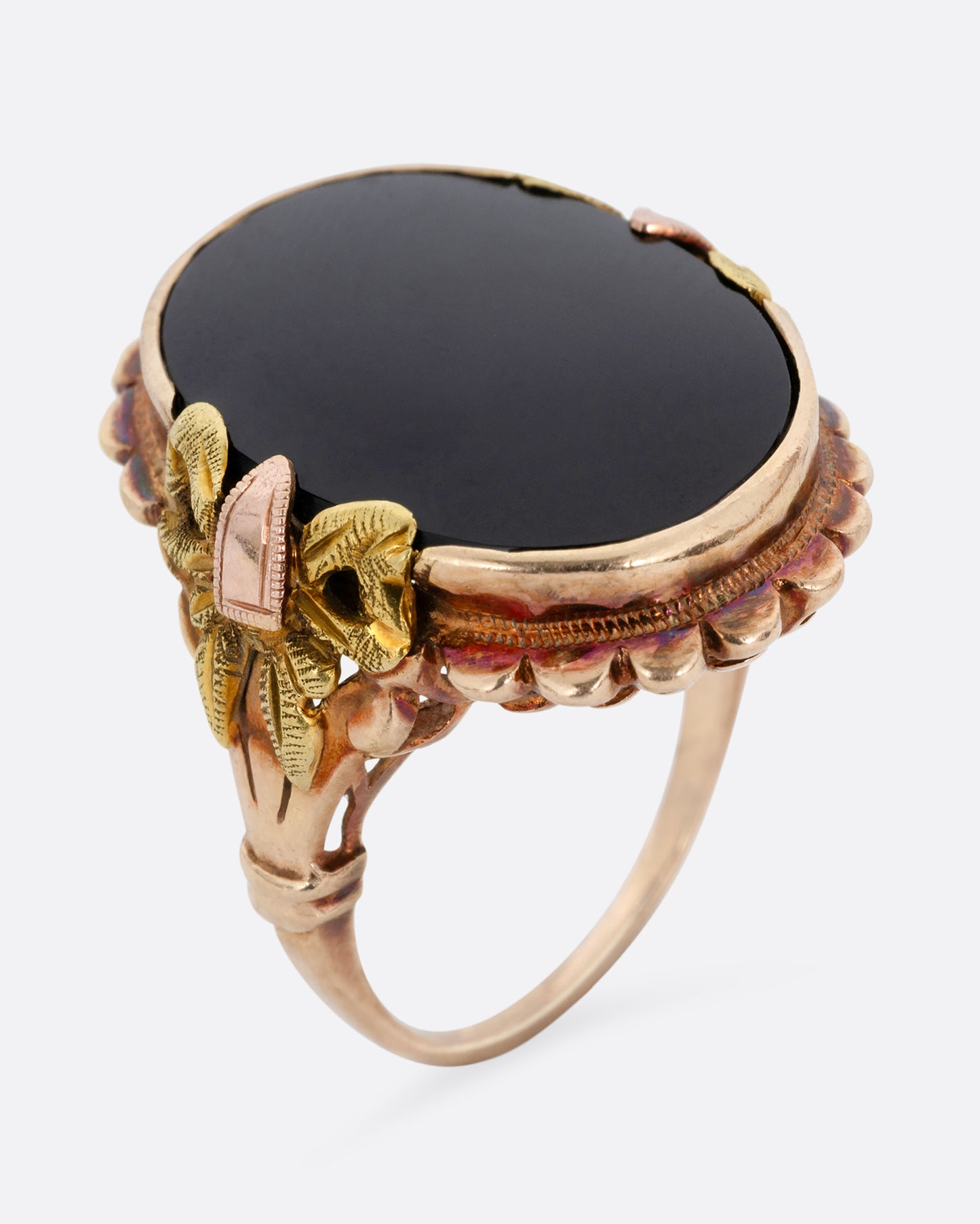 A yellow, rose, and green gold ring with a scalloped bezel, bow- topped shoulders, and an oval onyx. Shown from the side standing up.