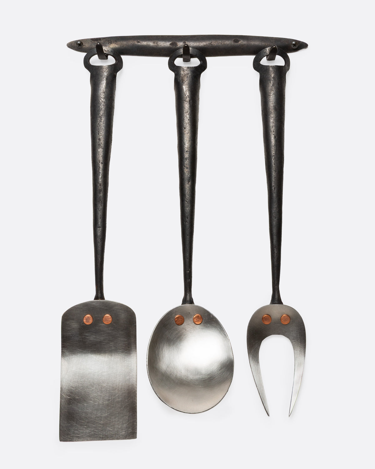 A set of stainless steel utensils with forged iron handles and a hanger for display. Shown from the front.