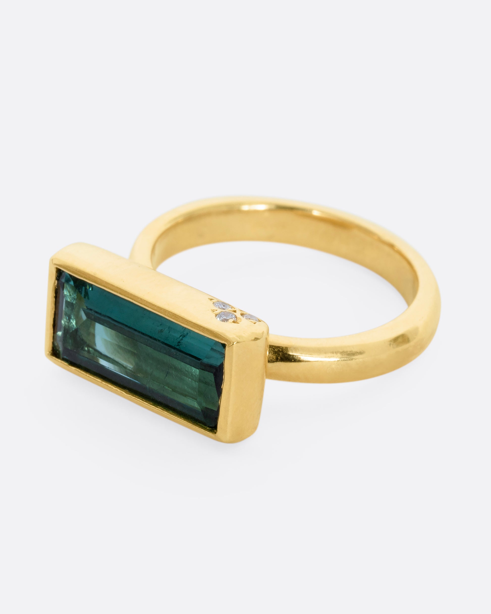 A yellow gold ring with a baguette indicolite tourmaline and small white diamonds. Shown from the side.