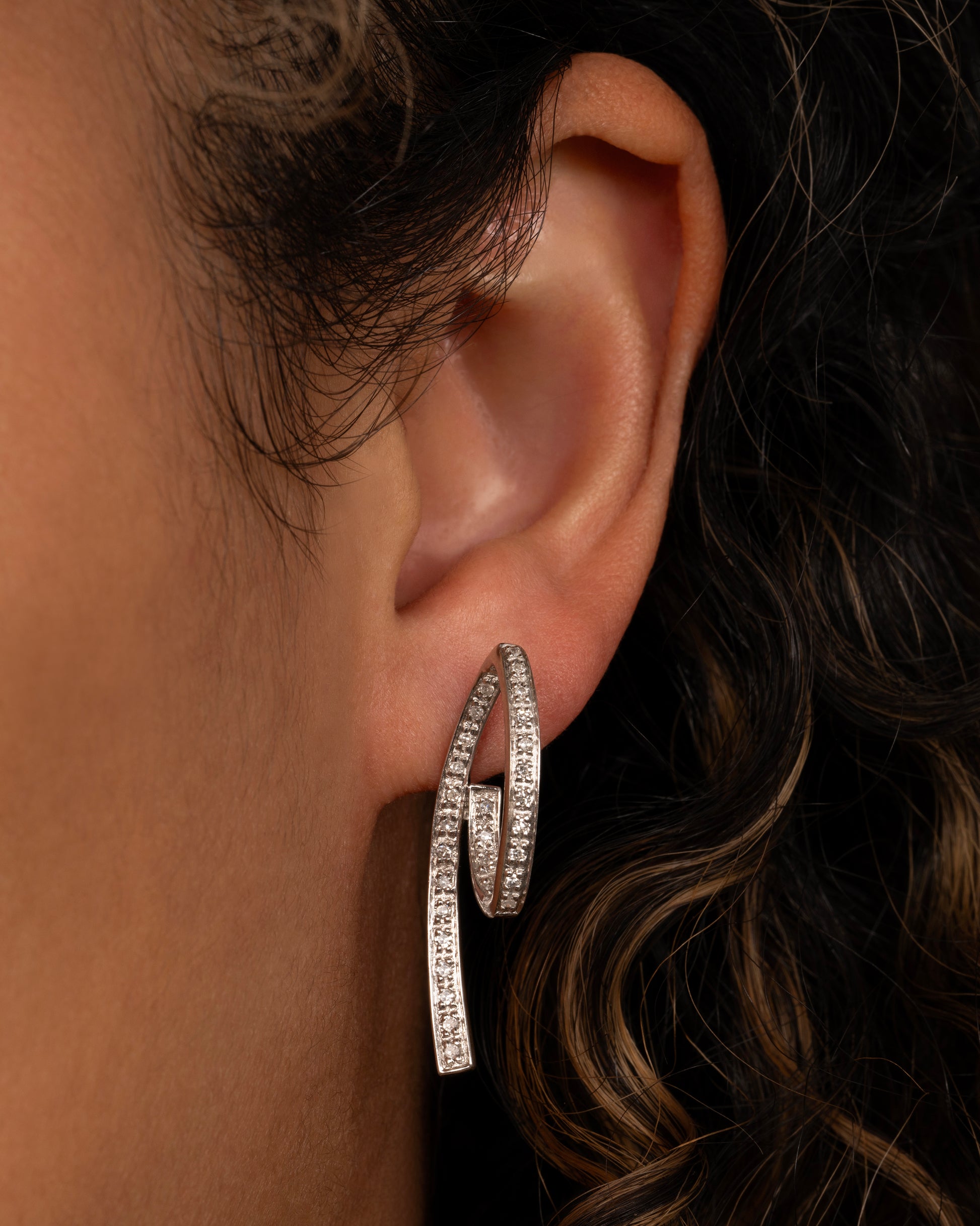 A pair of white gold, 3D loop de loop earrings, lined with diamonds. Shown on an ear.