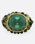 A yellow gold signet band with a hand carved winged hourglass in a bicolor green tourmaline. An array of green sapphire and tourmaline cabochons cover the band. Shown from the front.