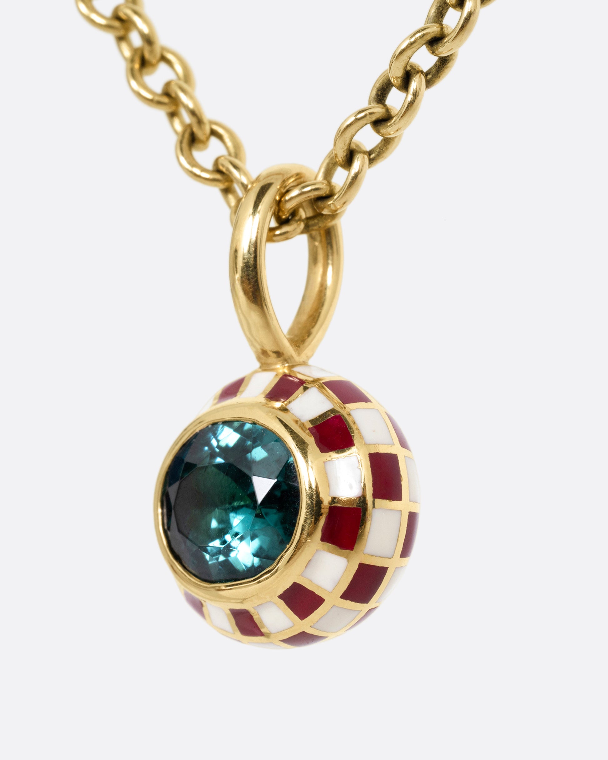 A yellow gold cable chain necklace with a red and cream enamel checkerboard pendant around a green tourmaline. The reverse side of the pendant has a checked pattern. Shown from the side.