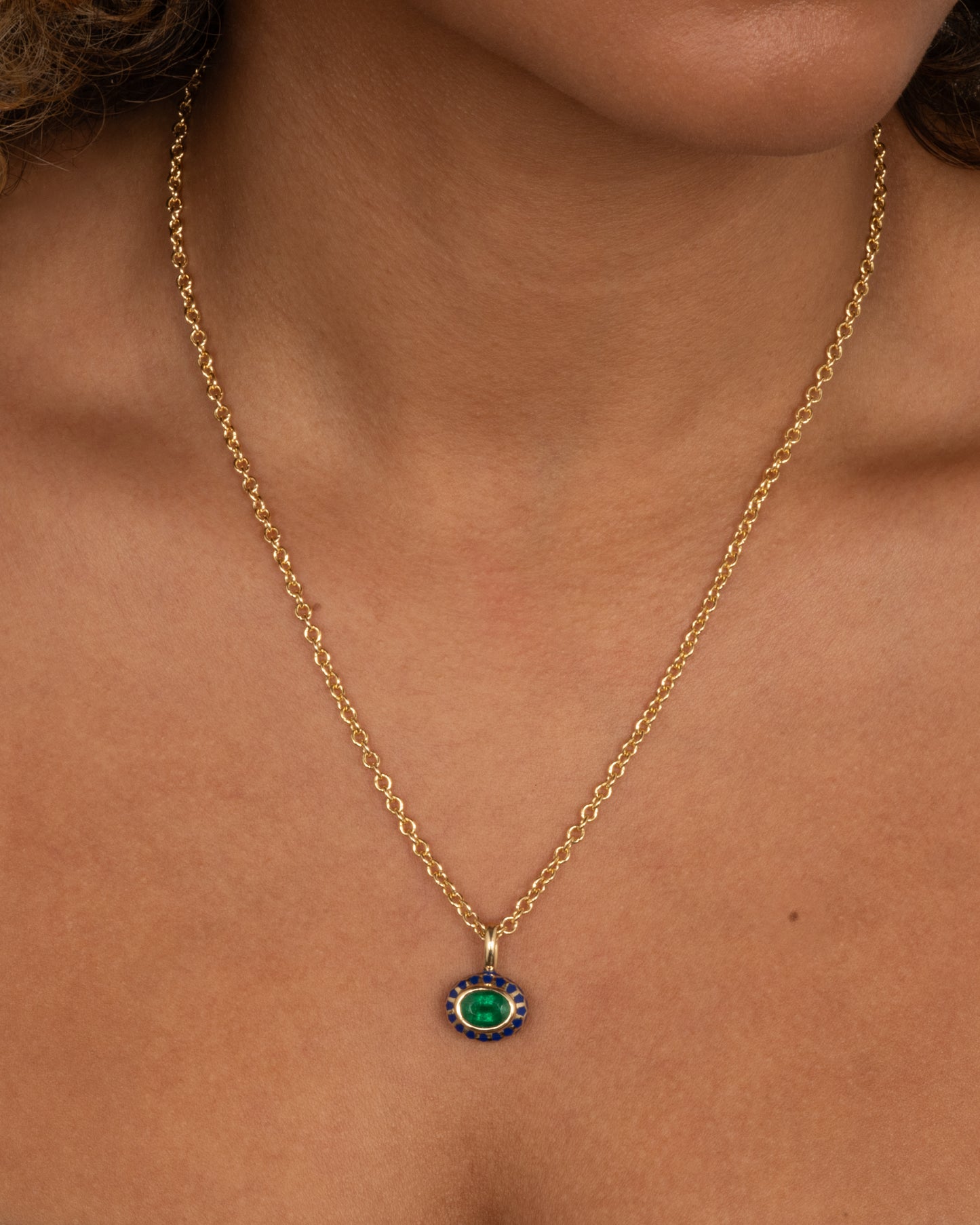 A yellow gold cable chain necklace with a pendant with a blue enamel pattern around an emerald. The pendant has a domed silhouette and floral enamel motif on its reverse. Shown on a person.