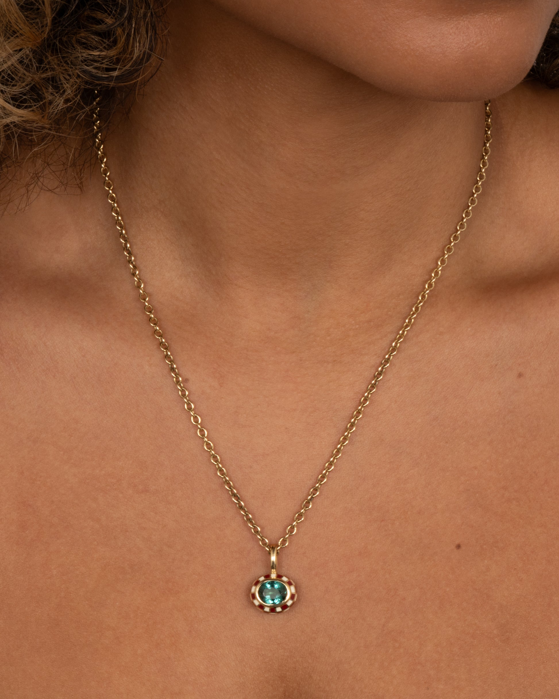 A yellow gold cable chain necklace with a red and cream enamel checkerboard pendant around a green tourmaline. The reverse side of the pendant has a checked pattern. Shown on a person.