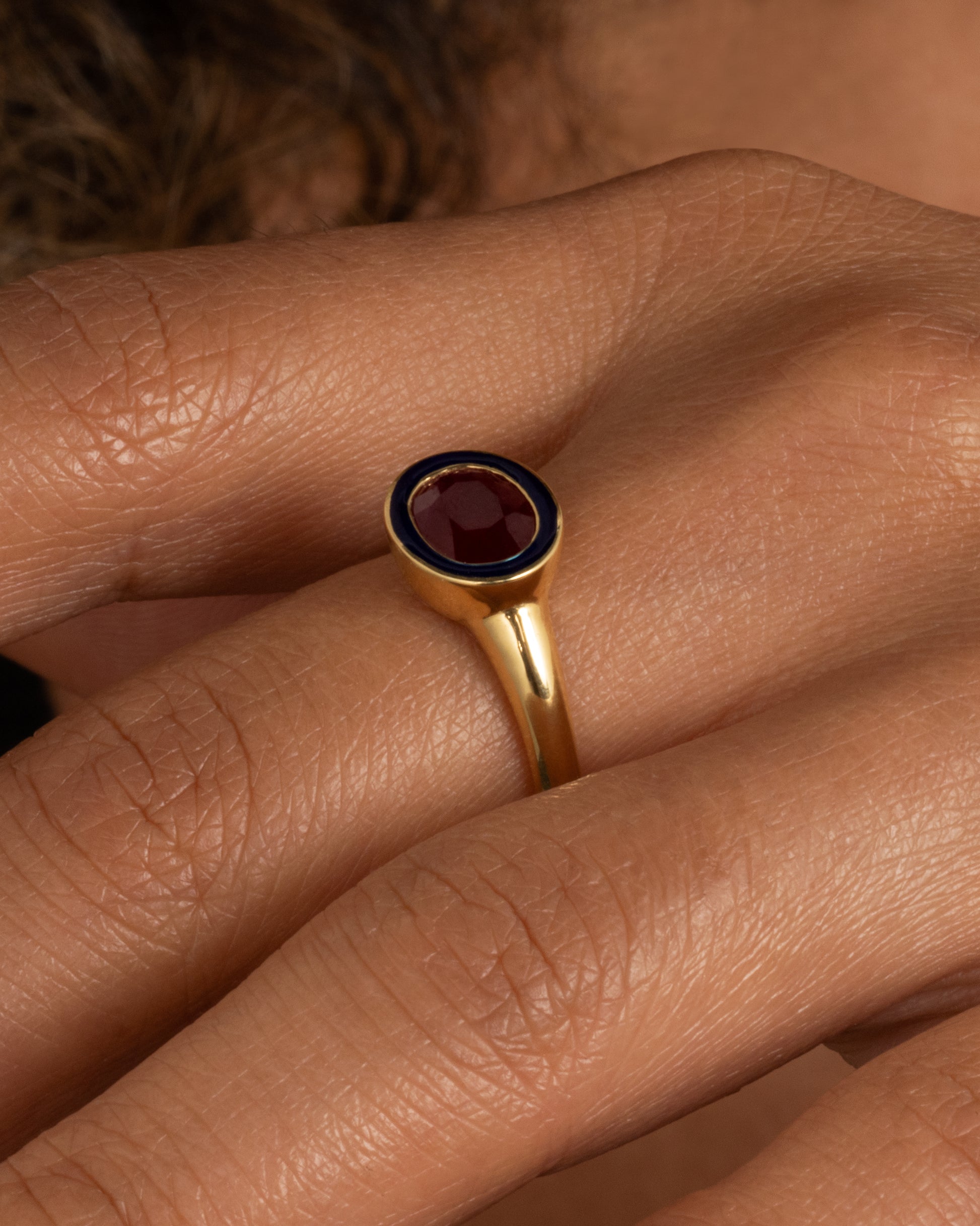 A yellow gold ring with a navy enamel and ruby center. Shown on a hand.