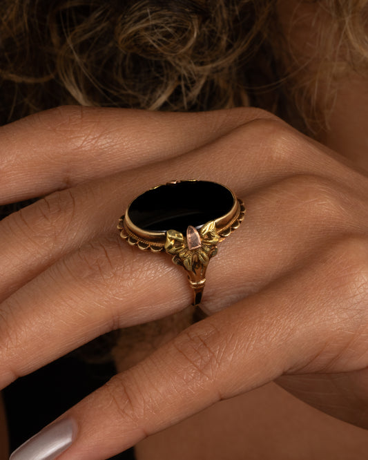 A yellow, rose, and green gold ring with a scalloped bezel, bow- topped shoulders, and an oval onyx. Shown worn