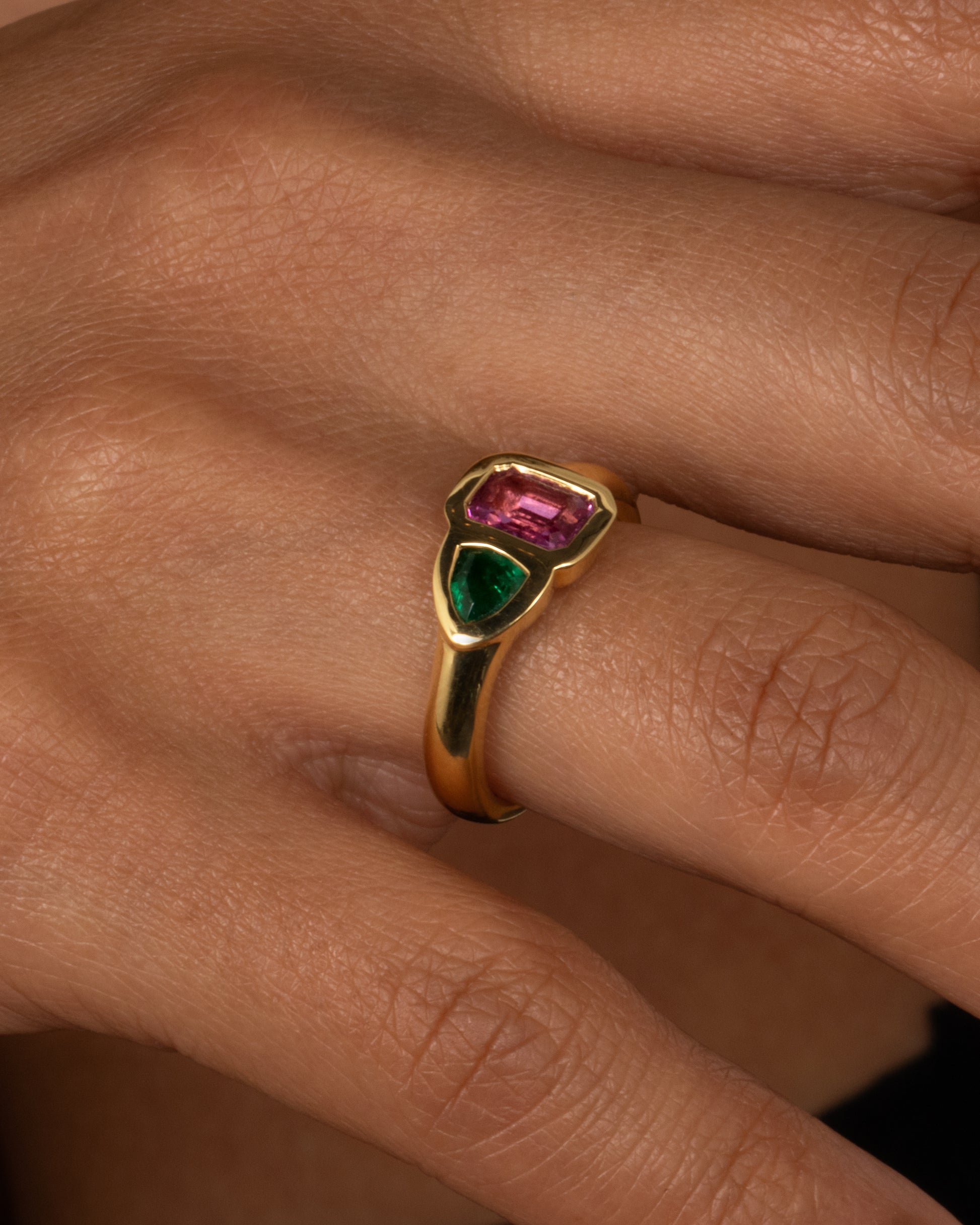 A yellow gold ring with an emerald and a pink sapphire, shown on a hand.