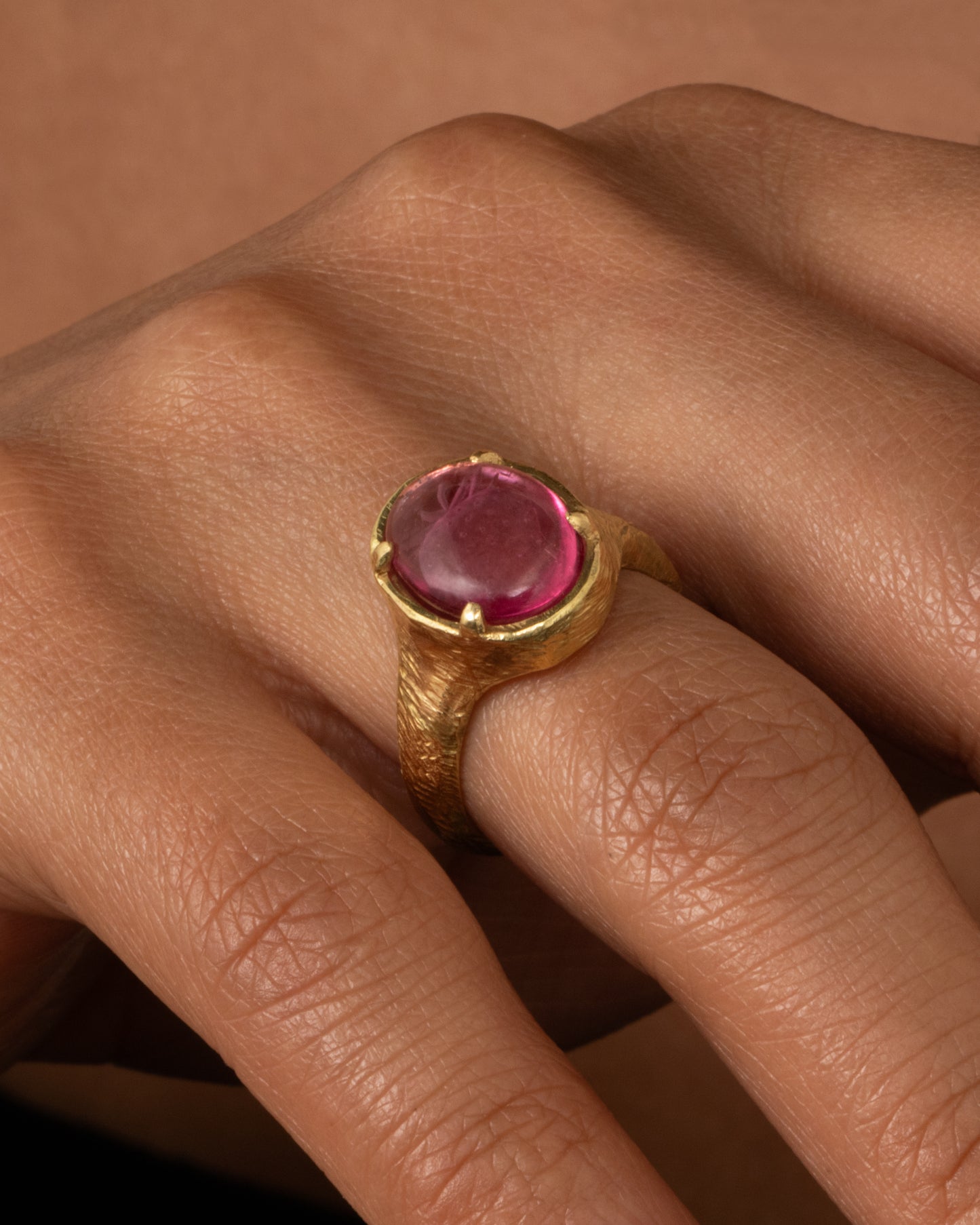 A yellow gold signet ring with a carved strawberry in a prong set pink garnet. Shown on a hand.