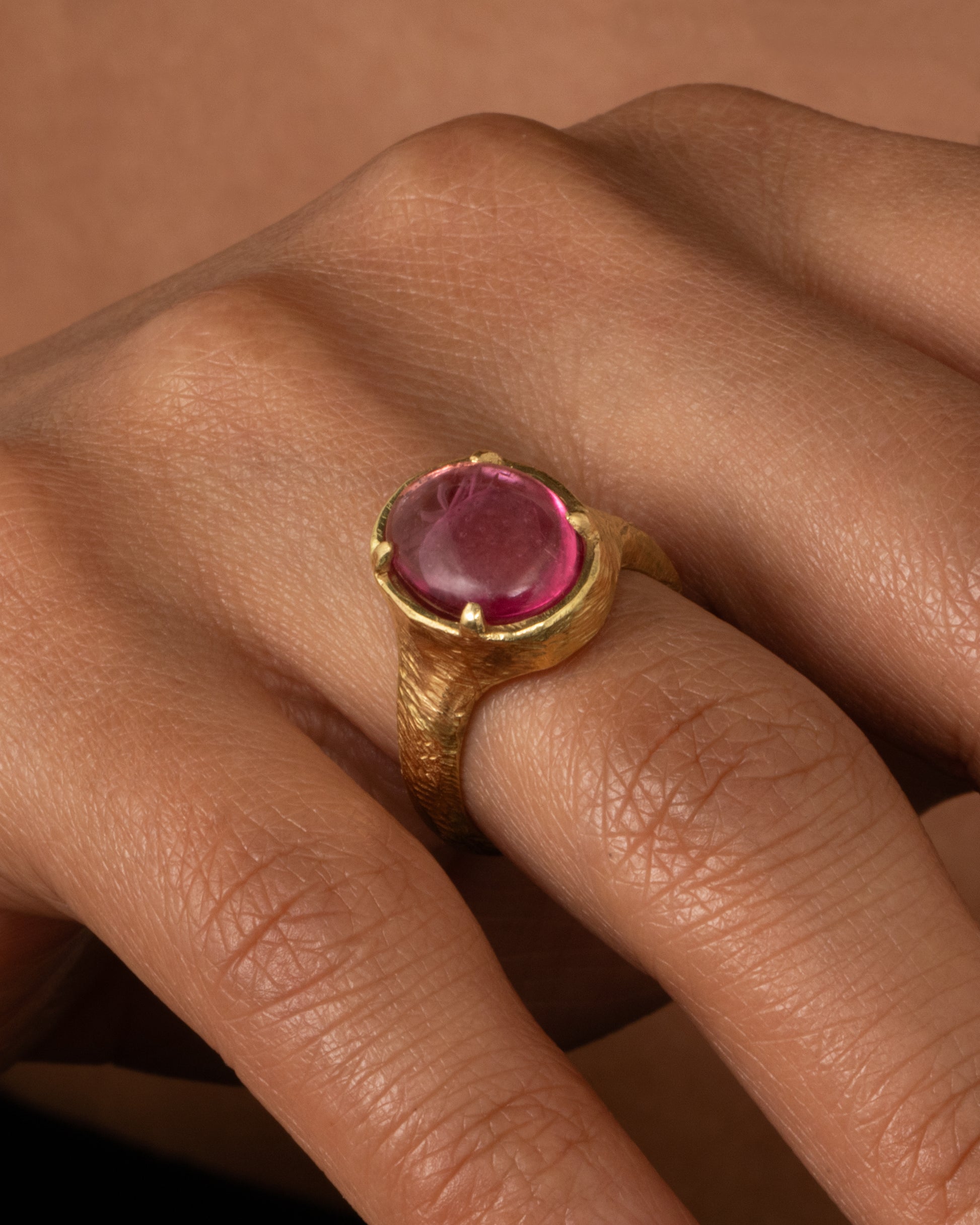 A yellow gold signet ring with a carved strawberry in a prong set pink garnet. Shown on a hand.