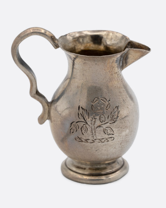 A tiny silver pitcher with an engraved flower, a spout, and handle. Shown from the side.