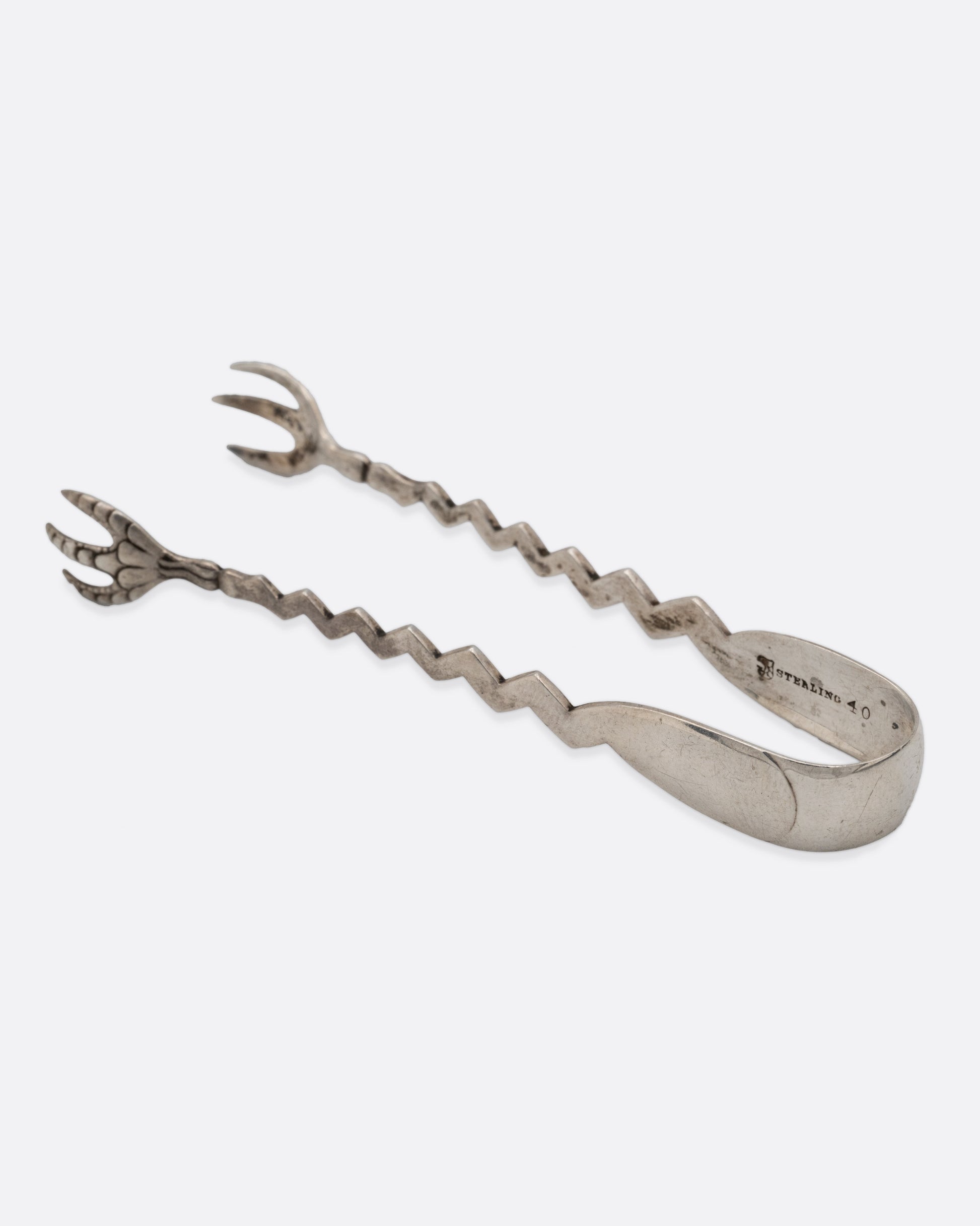 A pair of silver tongs with an arched top, zigzag arms, and claw like 'hands', shown from the side.