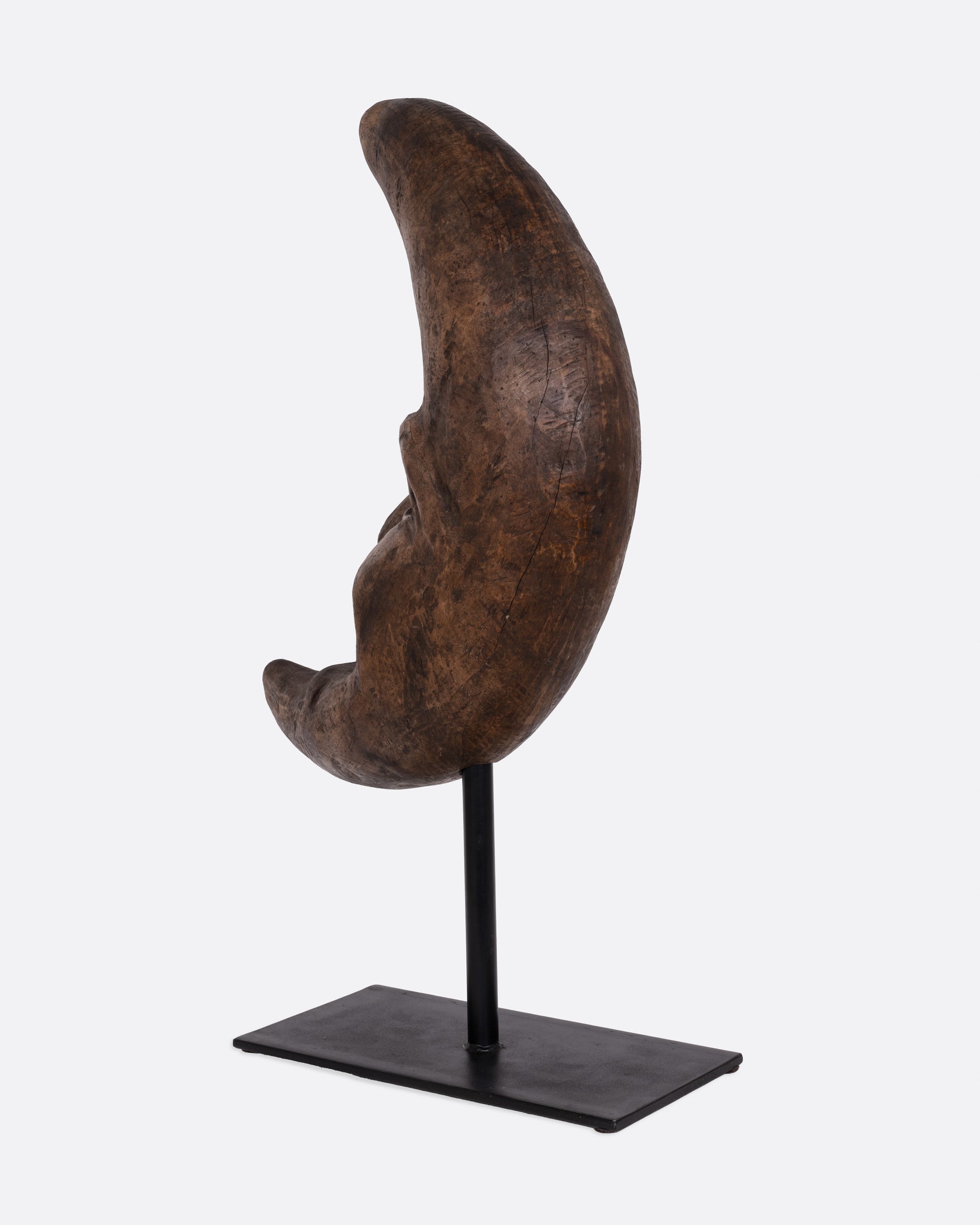 A wooden crescent moon takaan sculpture with a face on a black stand shown from the back.
