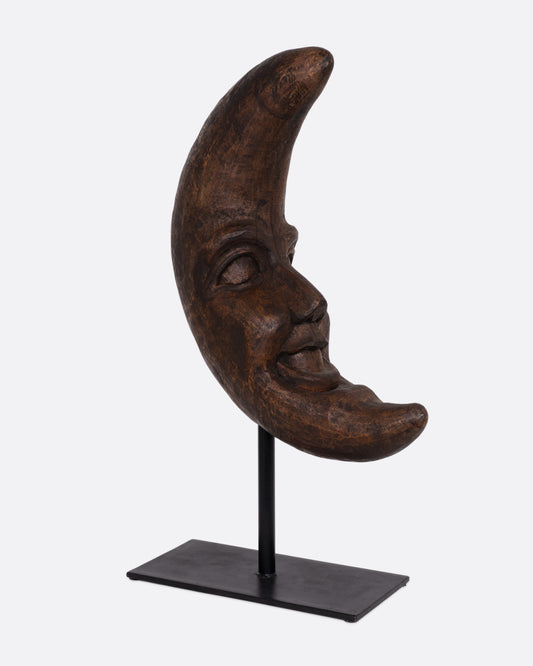 A wooden crescent moon takaan sculpture with a face on a black stand shown from the side.