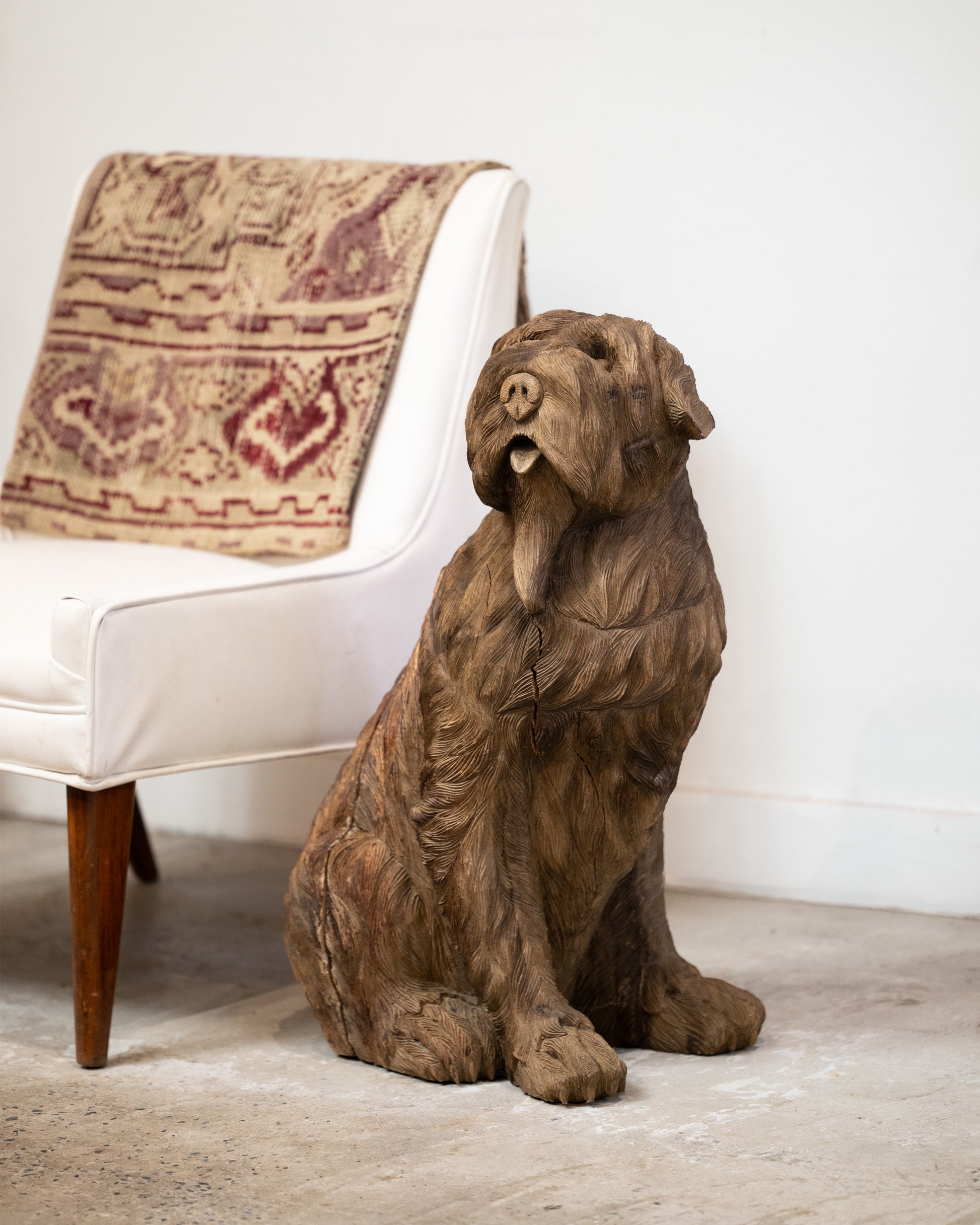 A wooden dog takaan sculpture shown from the front in front of a white chair.