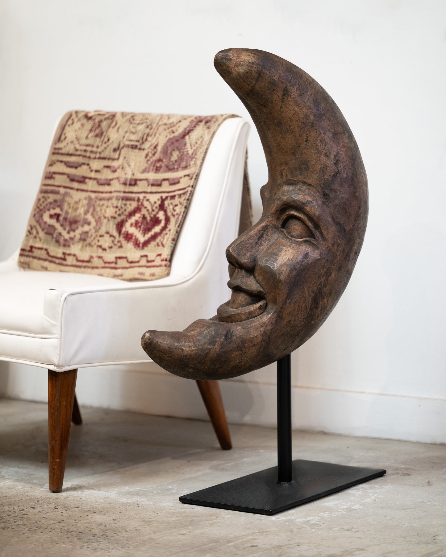 A wooden crescent moon takaan sculpture with a face on a black stand shown from the side in front of a white chair.