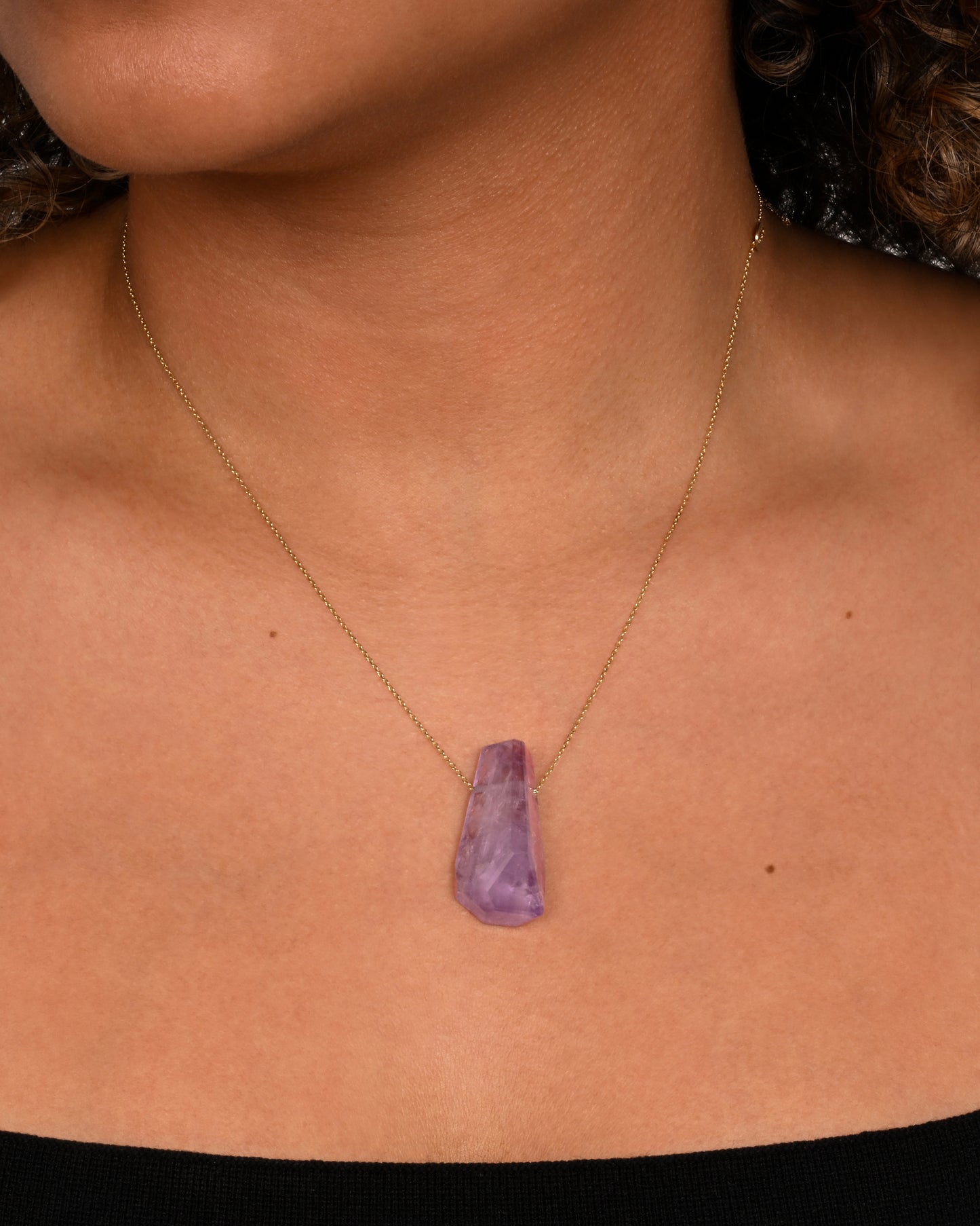 A geometric amethyst pendant hands from a dainty yellow gold chain. Shown worn around a neck.