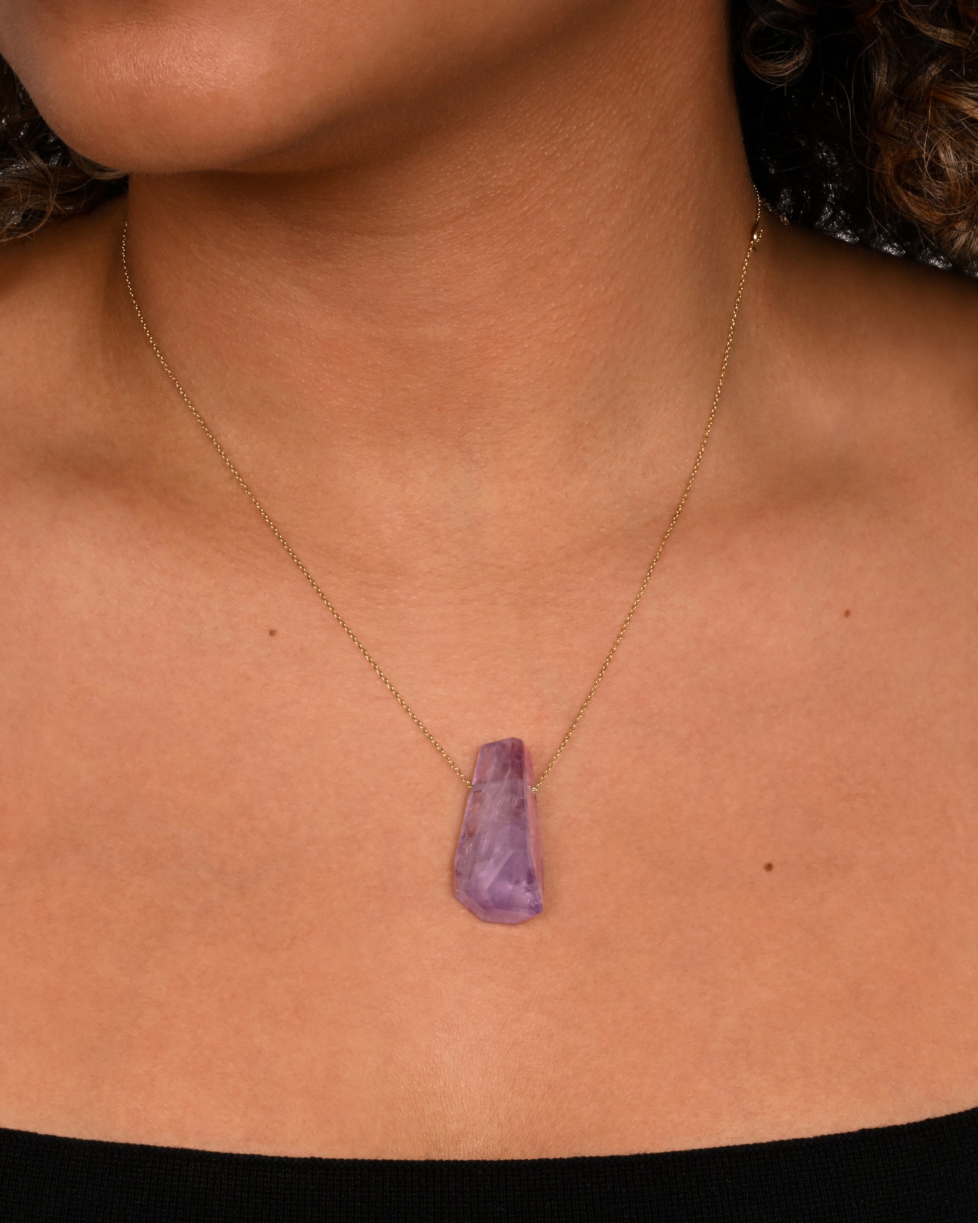 A geometric amethyst pendant hands from a dainty yellow gold chain. Shown worn around a neck.