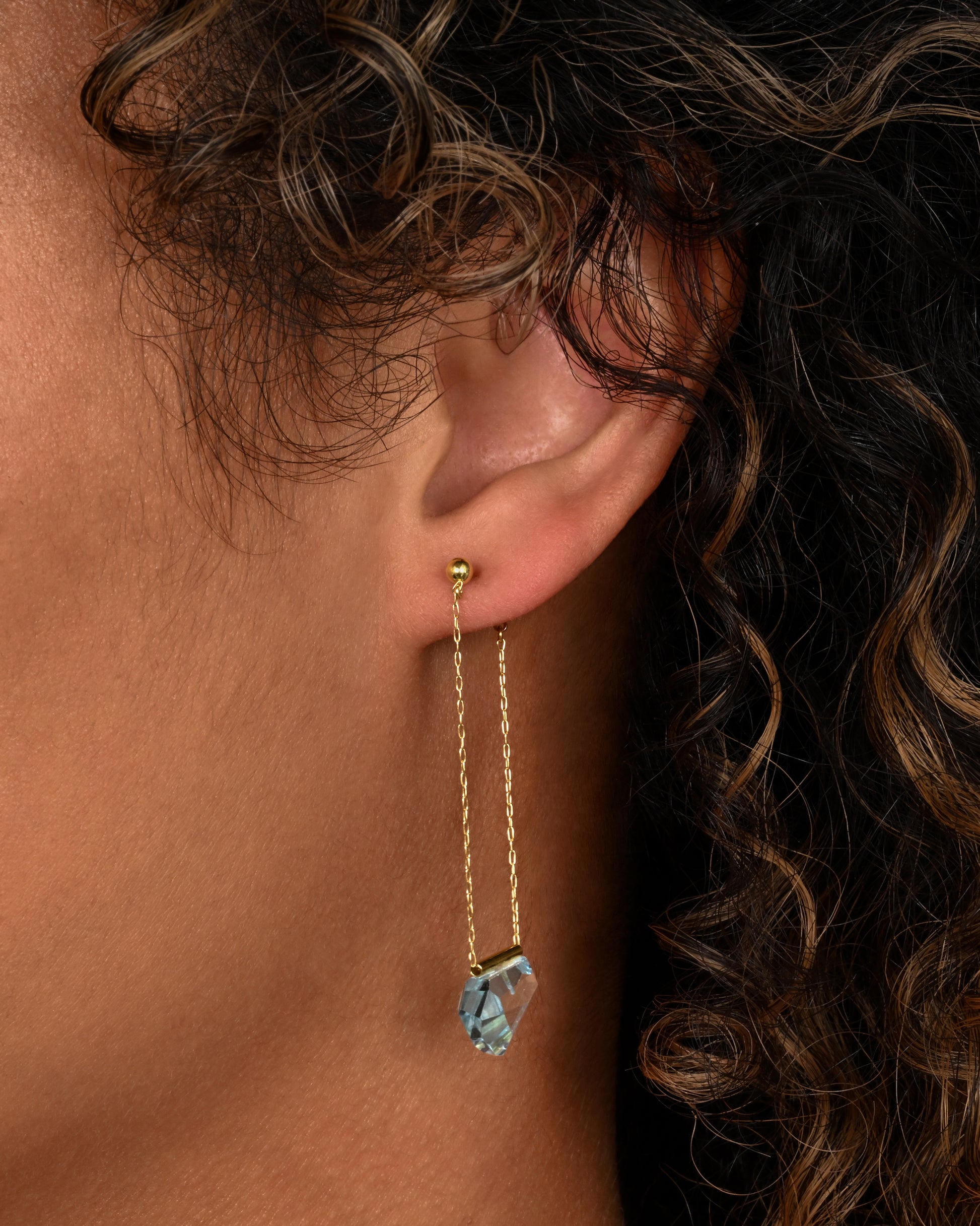 A pair of yellow gold chain earrings with free form blue topaz. Shown on an ear.