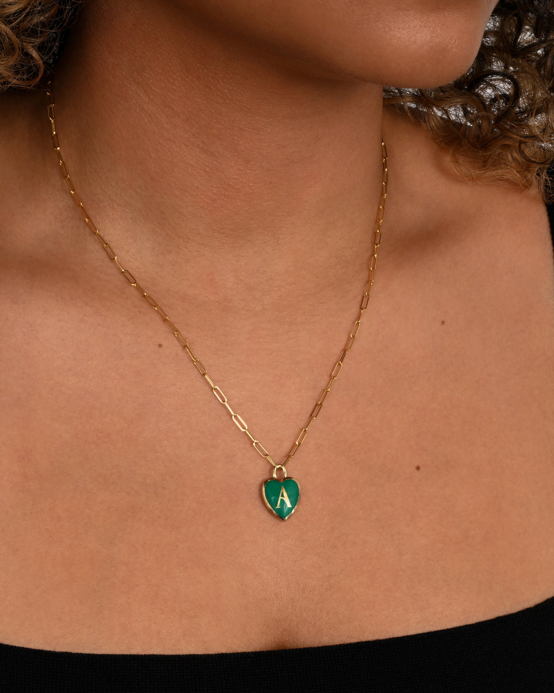 A yellow gold  paperclip chain necklace with a green enamel heart shaped A pendant. Shown from the front on a person.
