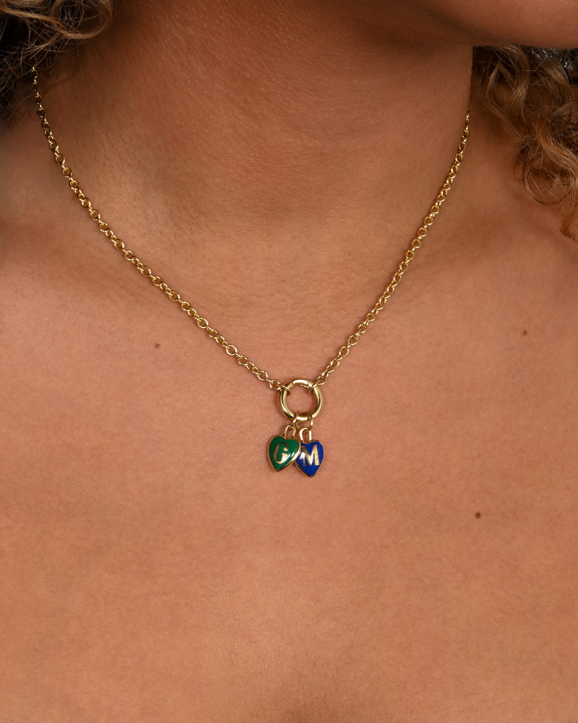 A yellow gold letter "B" heart shaped charm with blue enamel. A different letter version of the same charm style shown worn on a chain styled with another heart charm.