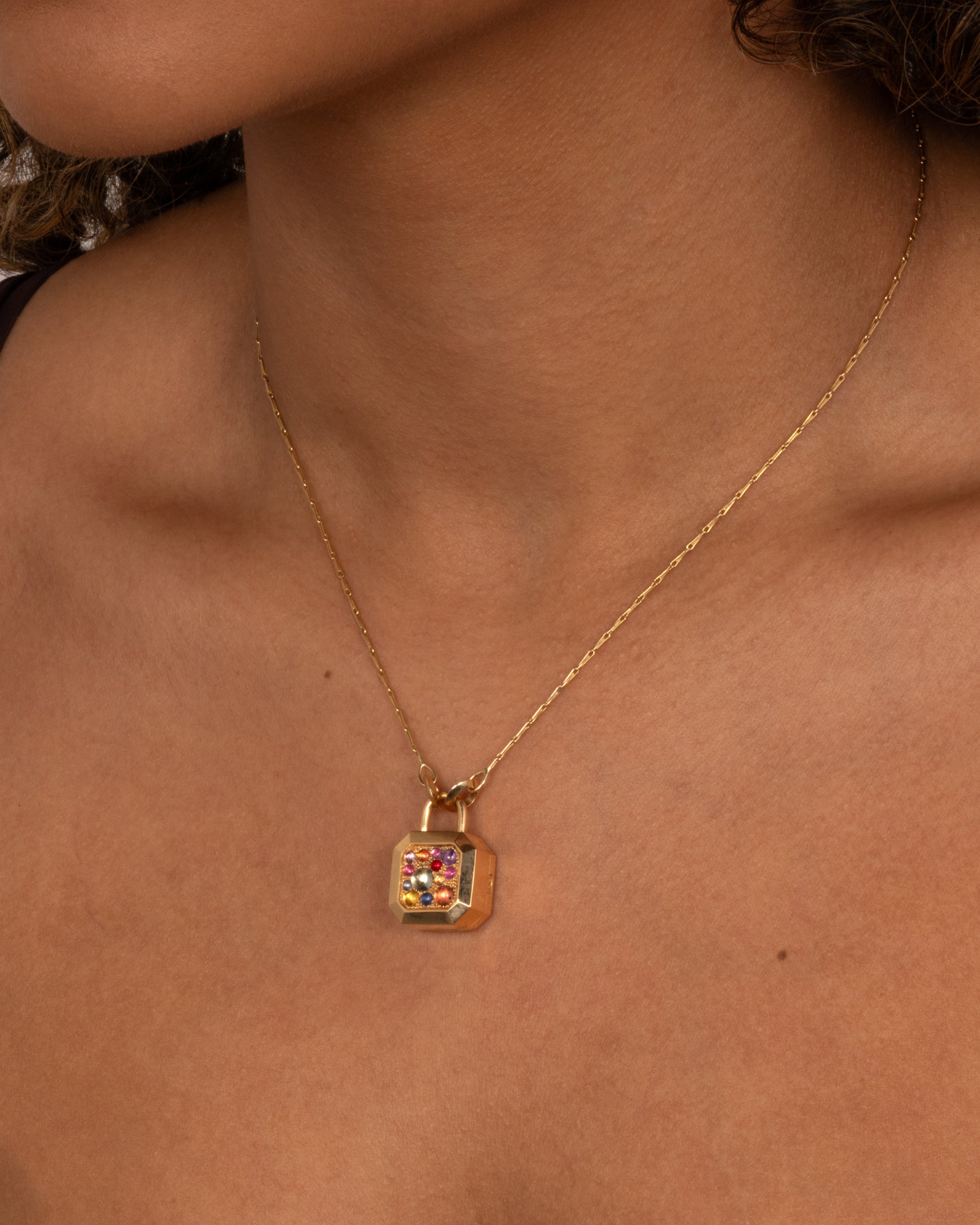 A yellow gold necklace with a padlock shaped locket pendant with multicolored sapphire on its face. Shown worn around the neck.
