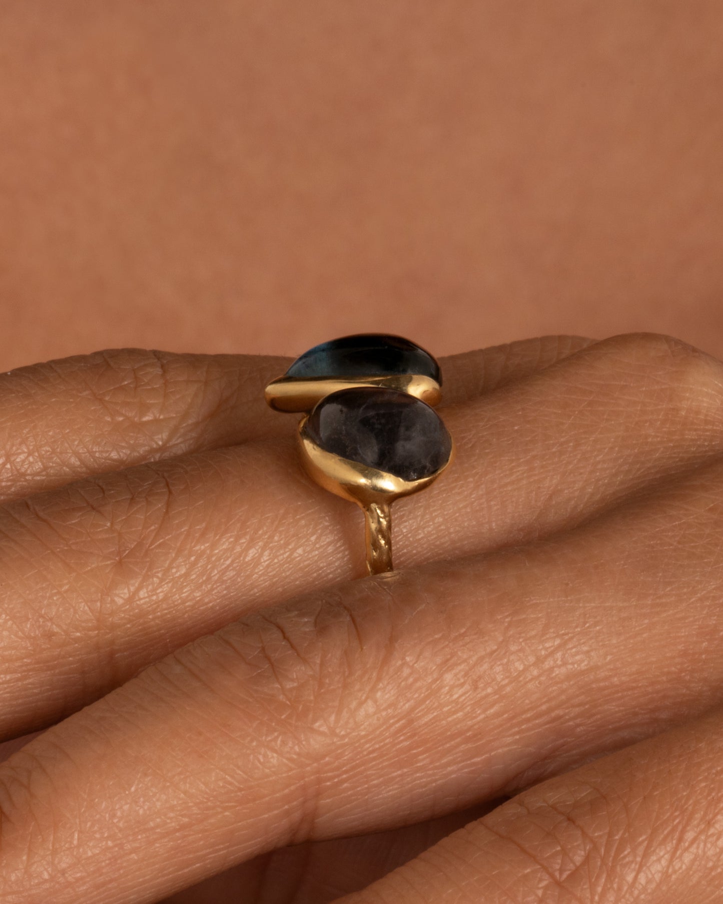 A yellow gold ring with a pair of blue green tourmalines and a textured hand- carved band, shown from the side on a hand.