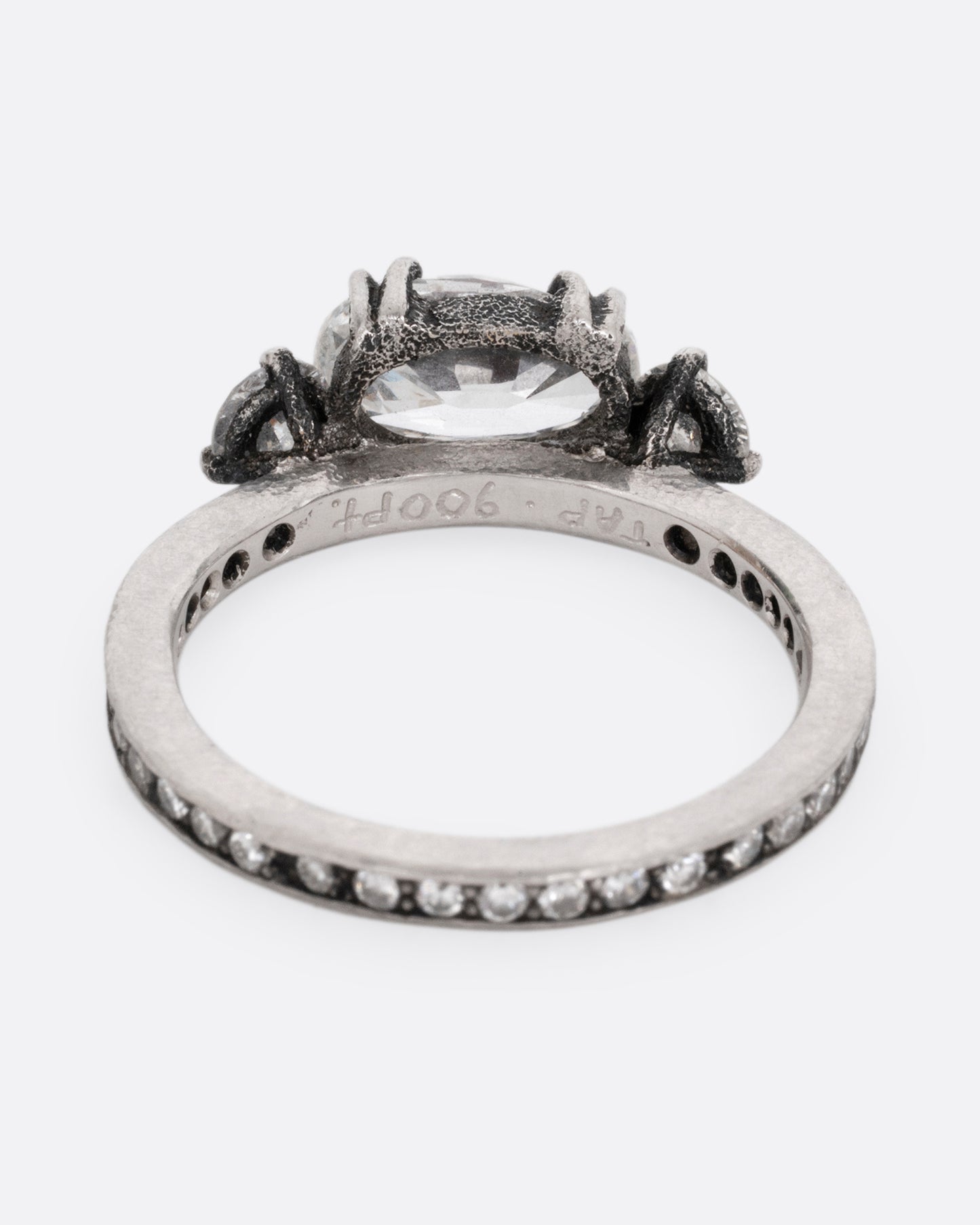 A platinum ring with a trio of diamonds, set upon a contrasting darkened channel of bead set diamonds. The elongated cushion cut center diamond is accented by two reverse set brilliant cut diamonds. Shown from the back.
