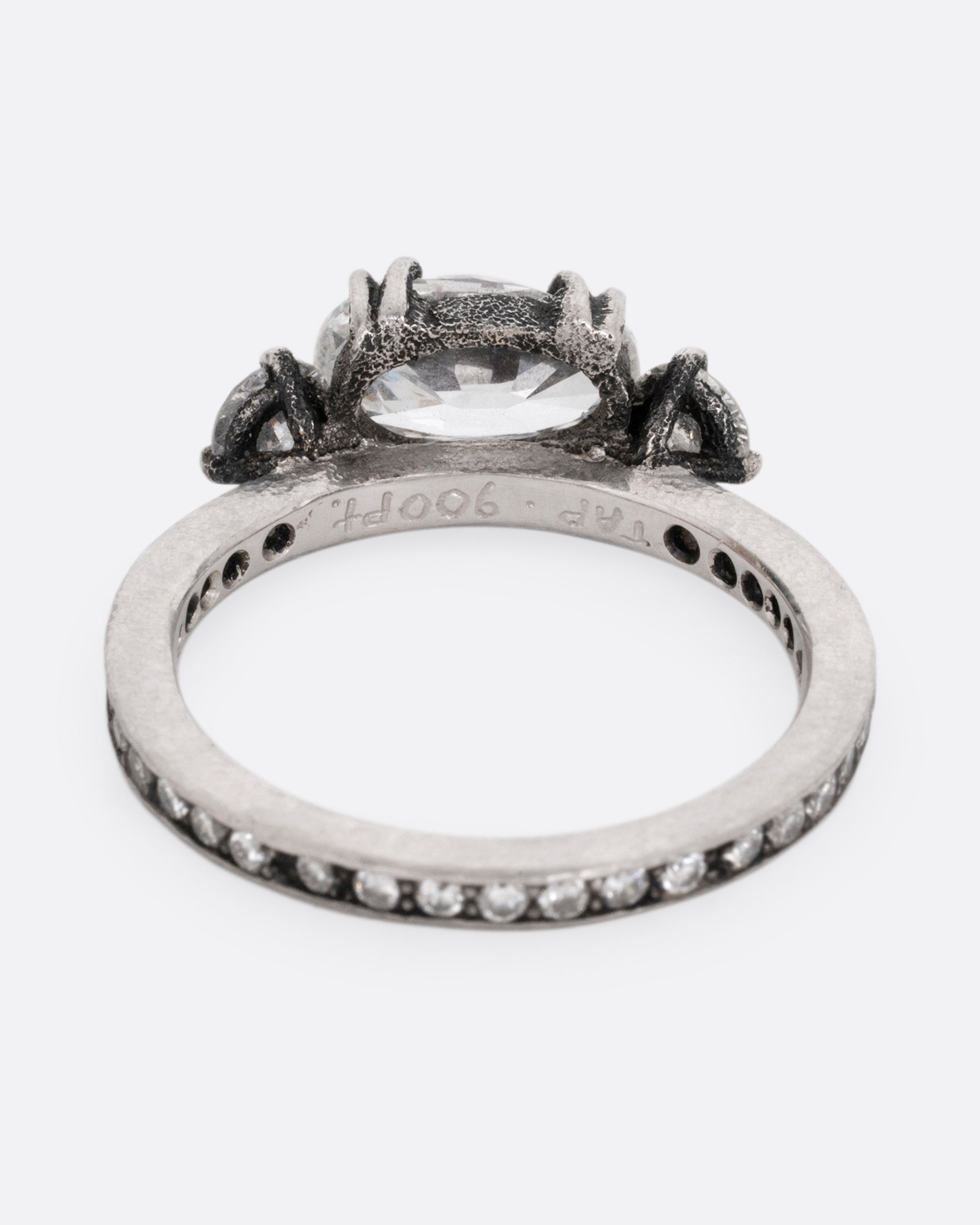 A platinum ring with a trio of diamonds, set upon a contrasting darkened channel of bead set diamonds. The elongated cushion cut center diamond is accented by two reverse set brilliant cut diamonds. Shown from the back.