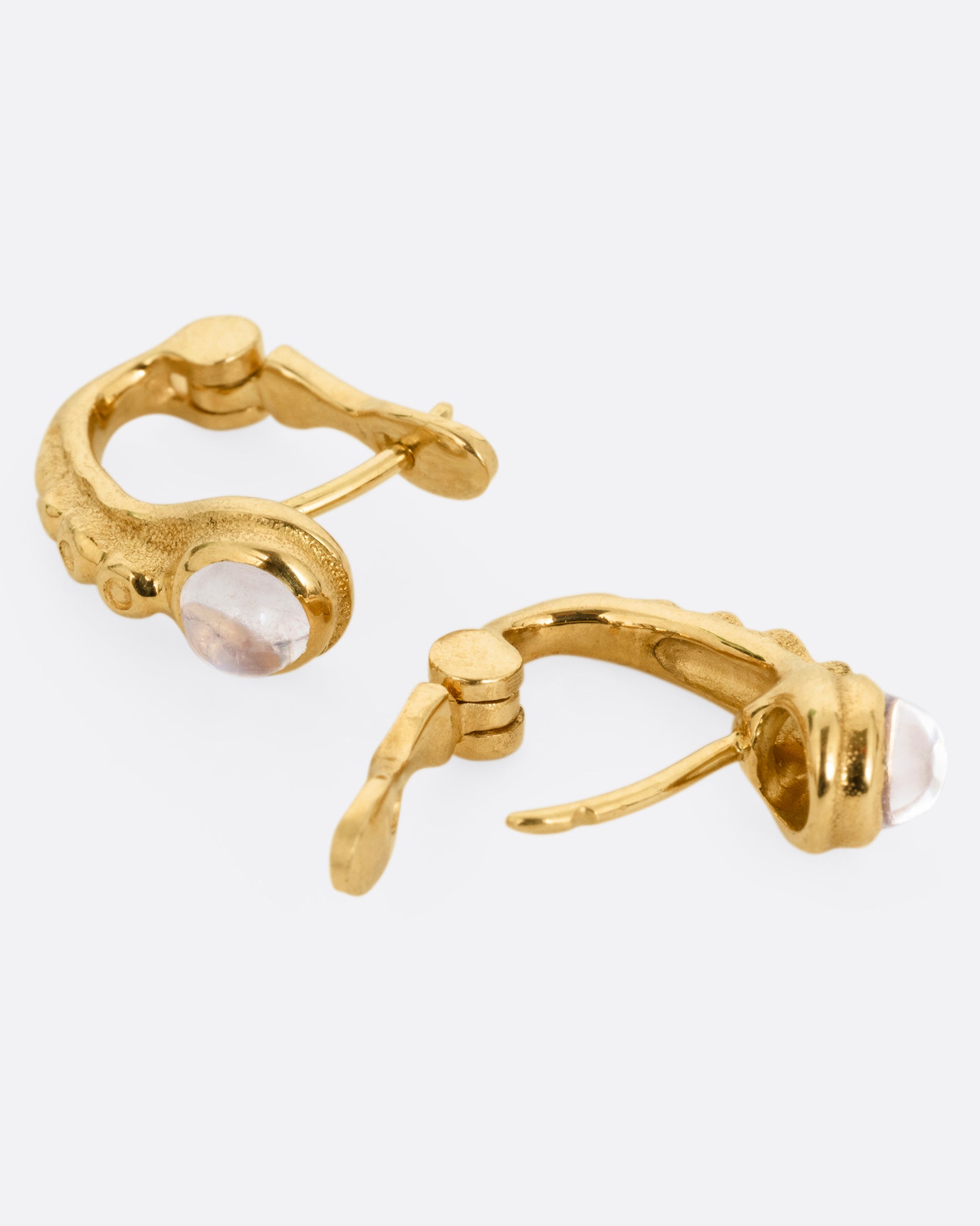 A pair of yellow gold hoop earrings with granulation and moonstones in each. Shown lying on their sides with one hoop open.