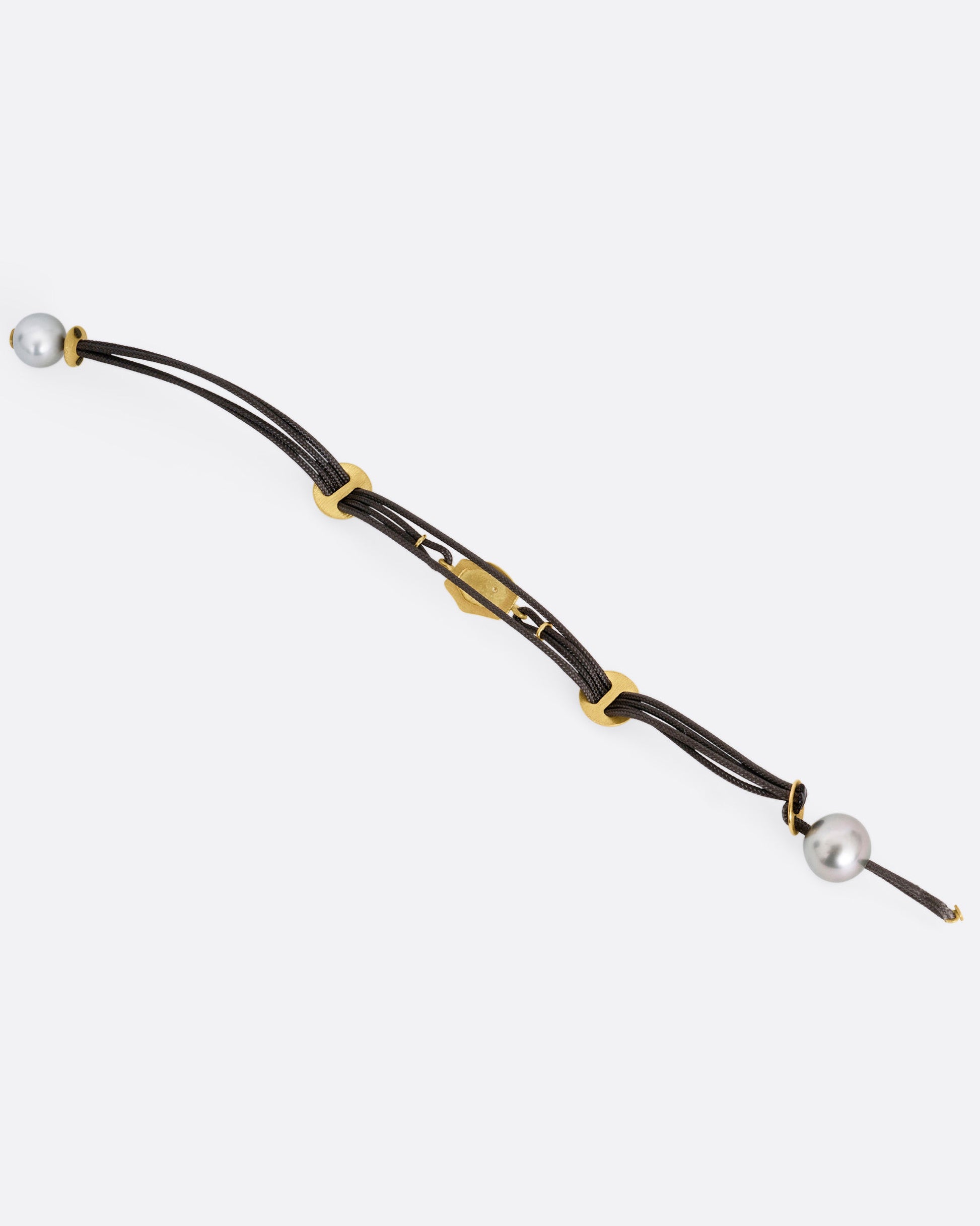 A  black cord bracelet with a diamond studded tag flanked by two gold slides and a pearl closure. Shown open from the back.