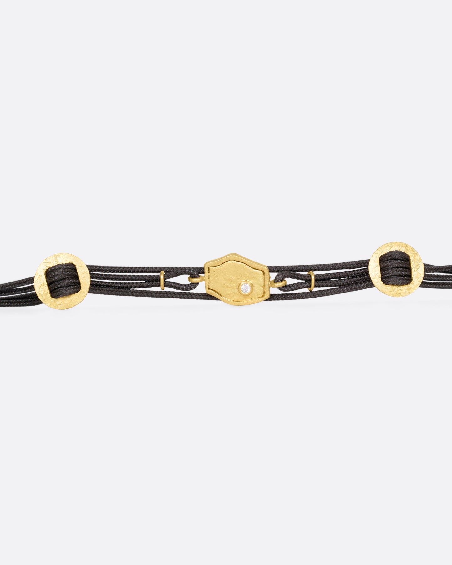 A  black cord bracelet with a diamond studded tag flanked by two gold slides and a pearl closure. Shown open from the front.