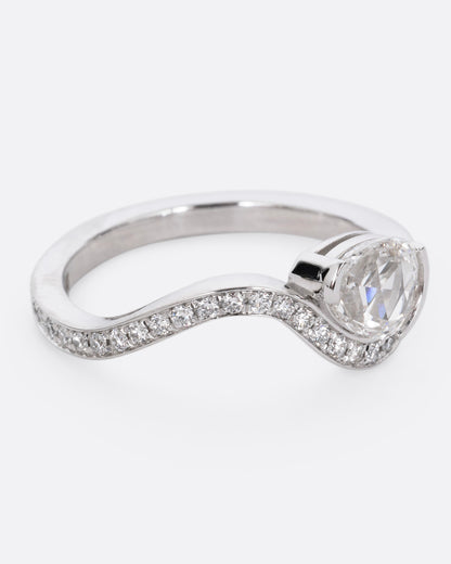 A diamond studded platinum ring with a wavy silhouette and a pear shaped rose cut diamond, shown from the side. 