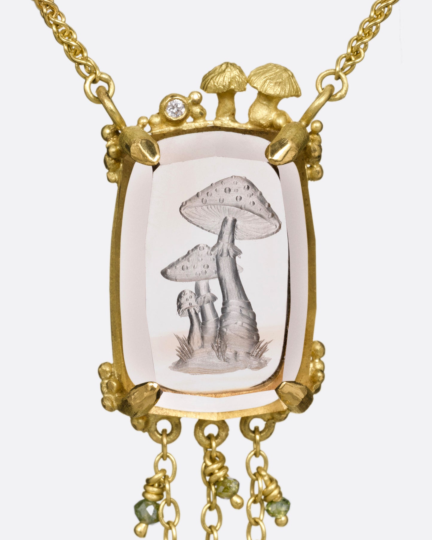 A yellow gold pendant necklace with a hand carved cluster of mushrooms in a light pink topaz with gold mushrooms and a diamond at the top edge. Three gold chains with green diamond beads hand from its base. Shown from the front.