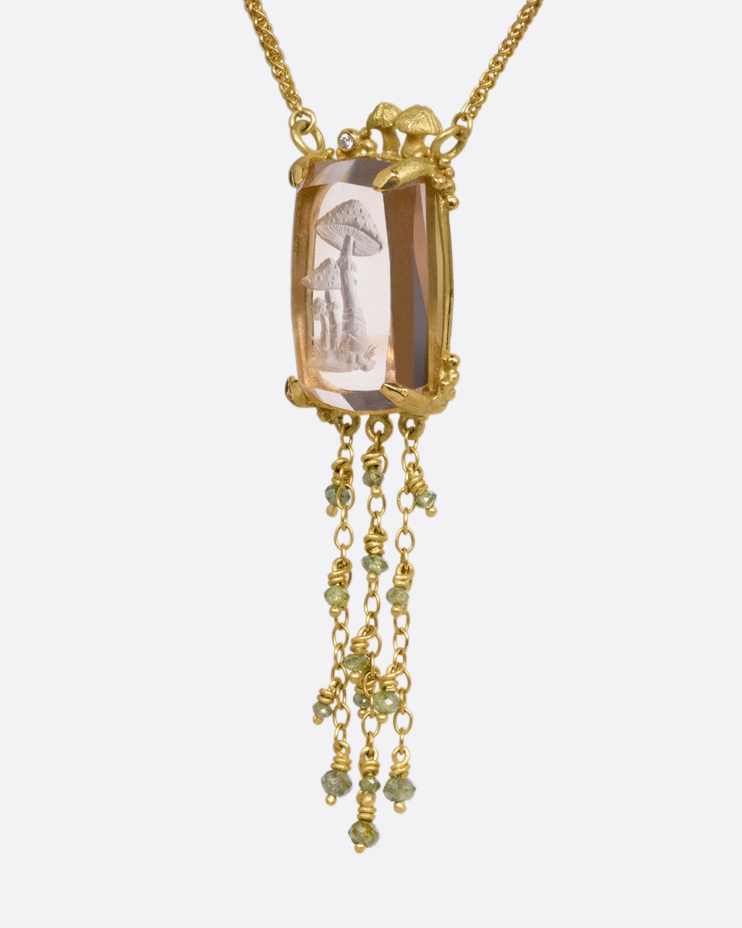 A yellow gold pendant necklace with a hand carved cluster of mushrooms in a light pink topaz with gold mushrooms and a diamond at the top edge. Three gold chains with green diamond beads hand from its base. Shown from the side.